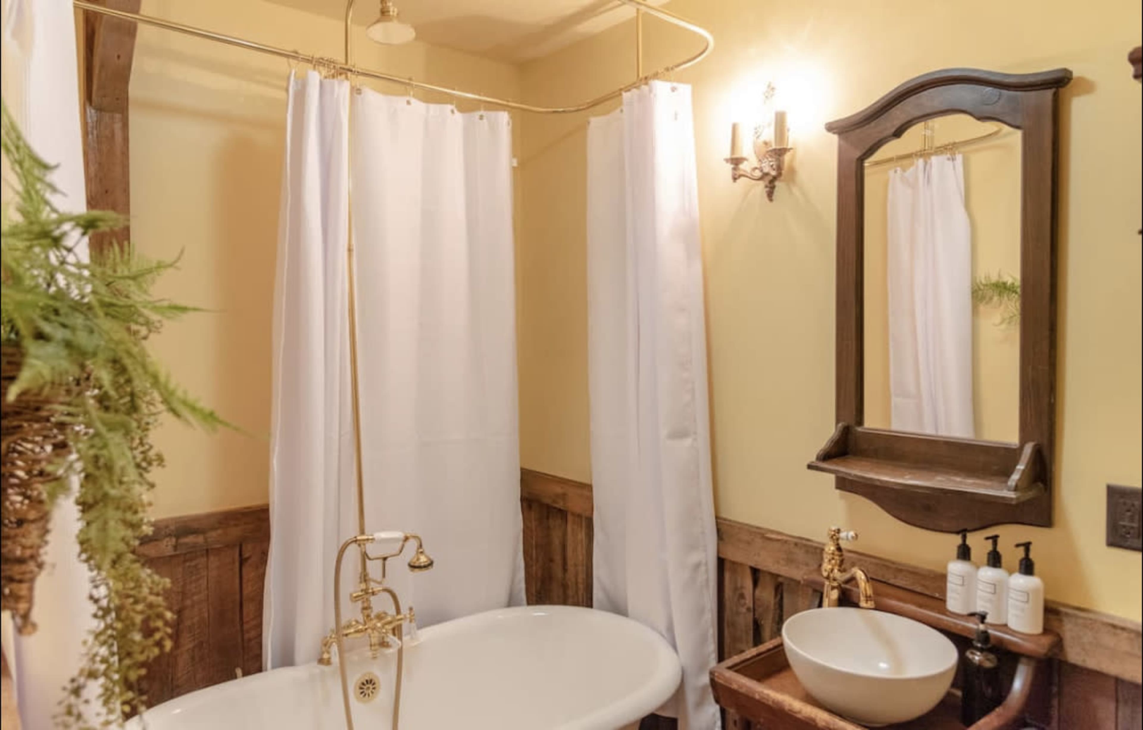 A bathroom featuring a freestanding bathtub, white shower curtains, a wooden accent wall, and a wall-mounted mirror with a small shelf.