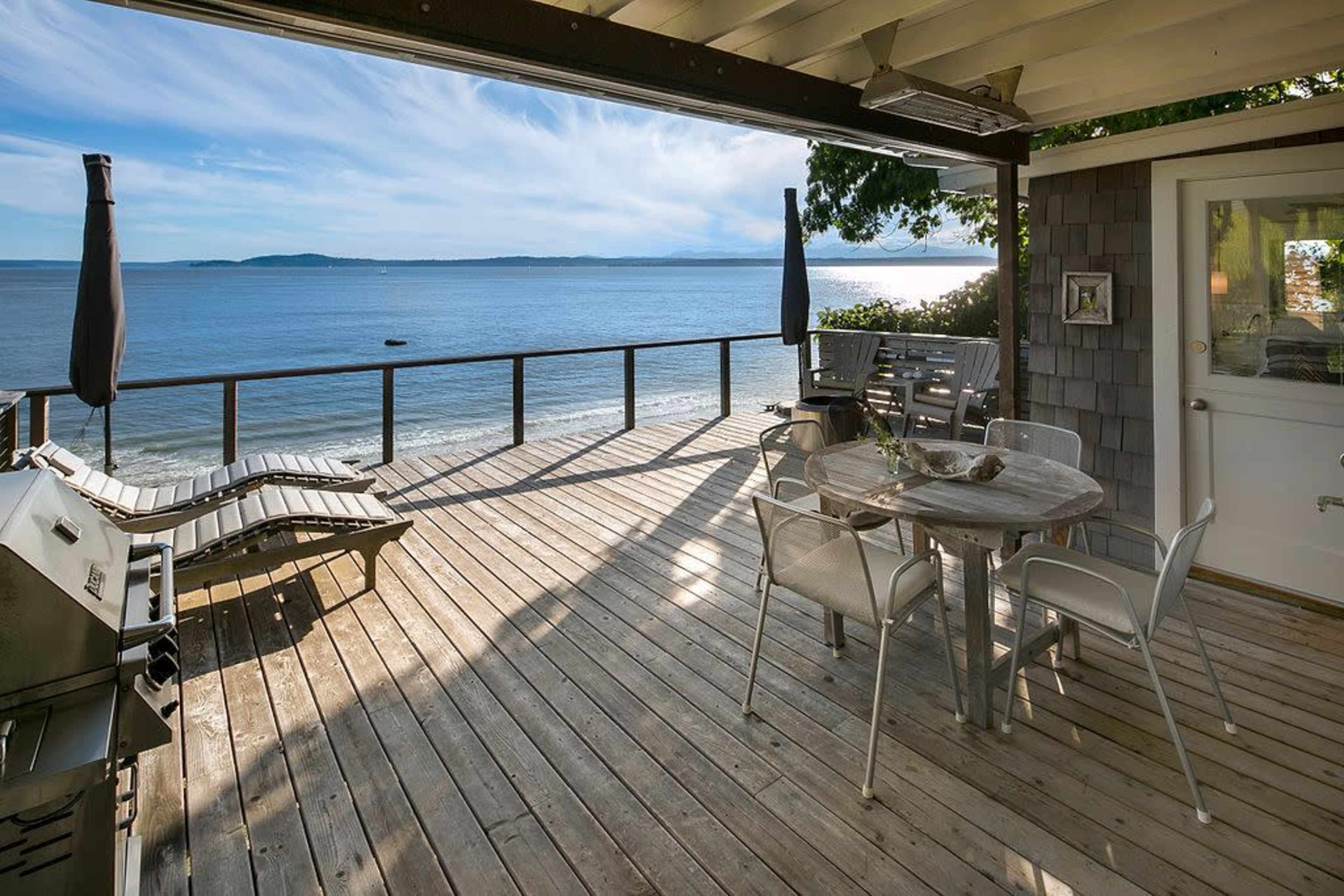 A wooden deck overlooks a calm body of water with lounge chairs and a dining table set for outdoor meals.