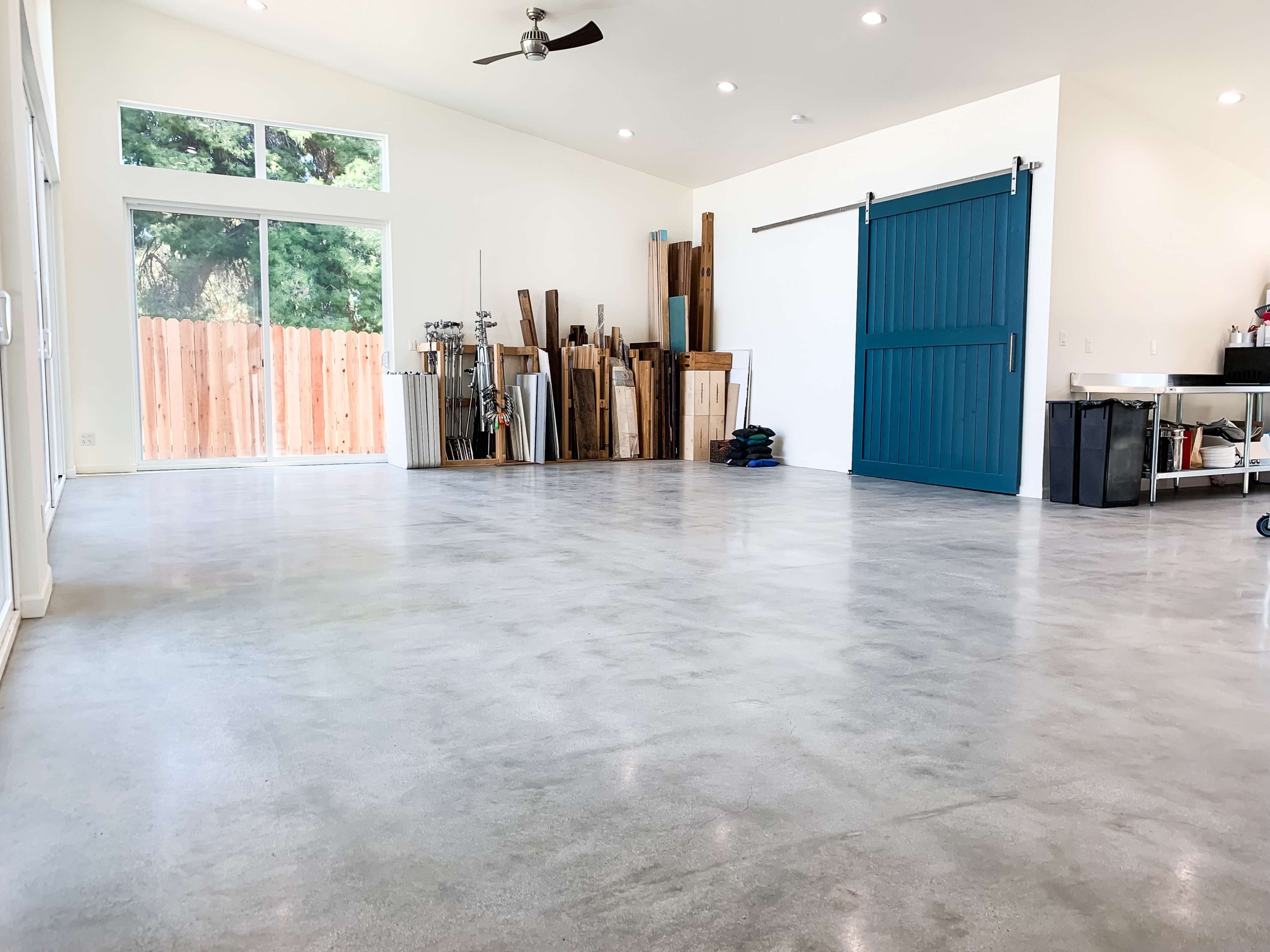 A spacious, minimalistic interior with polished concrete flooring, a large blue sliding barn door, and a stack of wooden planks along the wall.