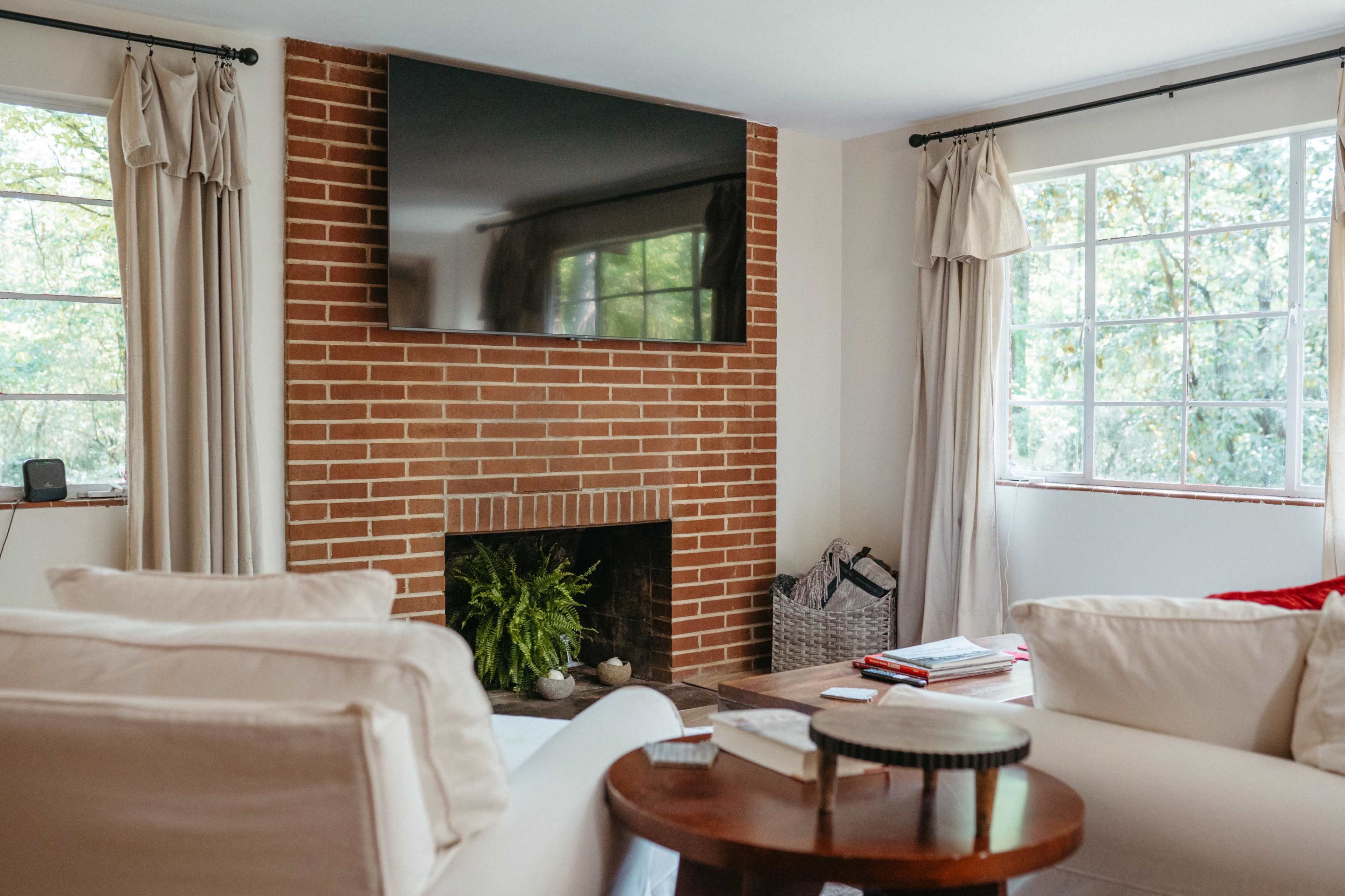 A living room features a brick fireplace underneath a mounted flat-screen television, with large windows allowing natural light and soft furnishings arranged around a small table.