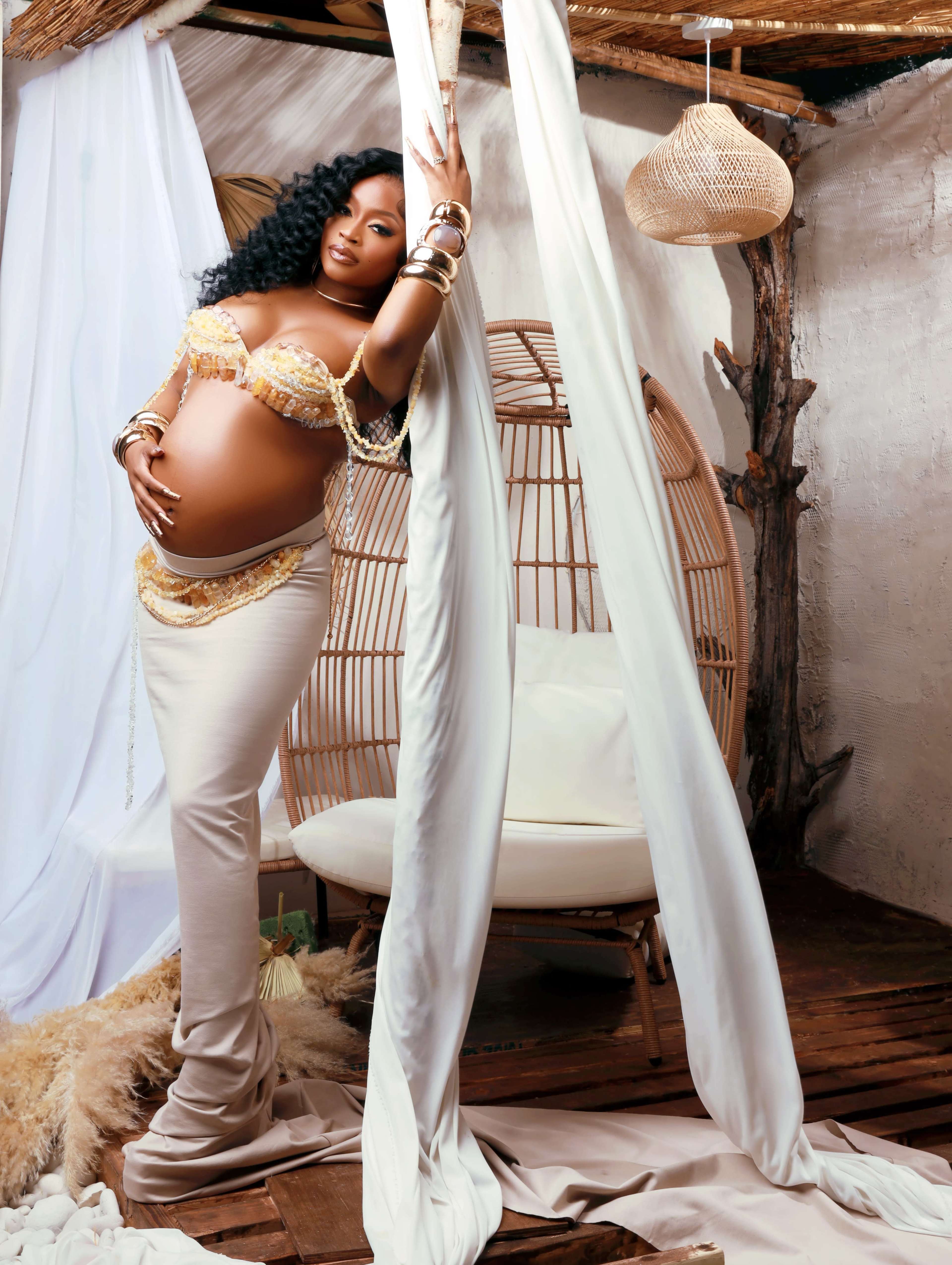 A pregnant woman in a form-fitting dress poses gracefully in a softly lit room adorned with natural textures and fabrics.