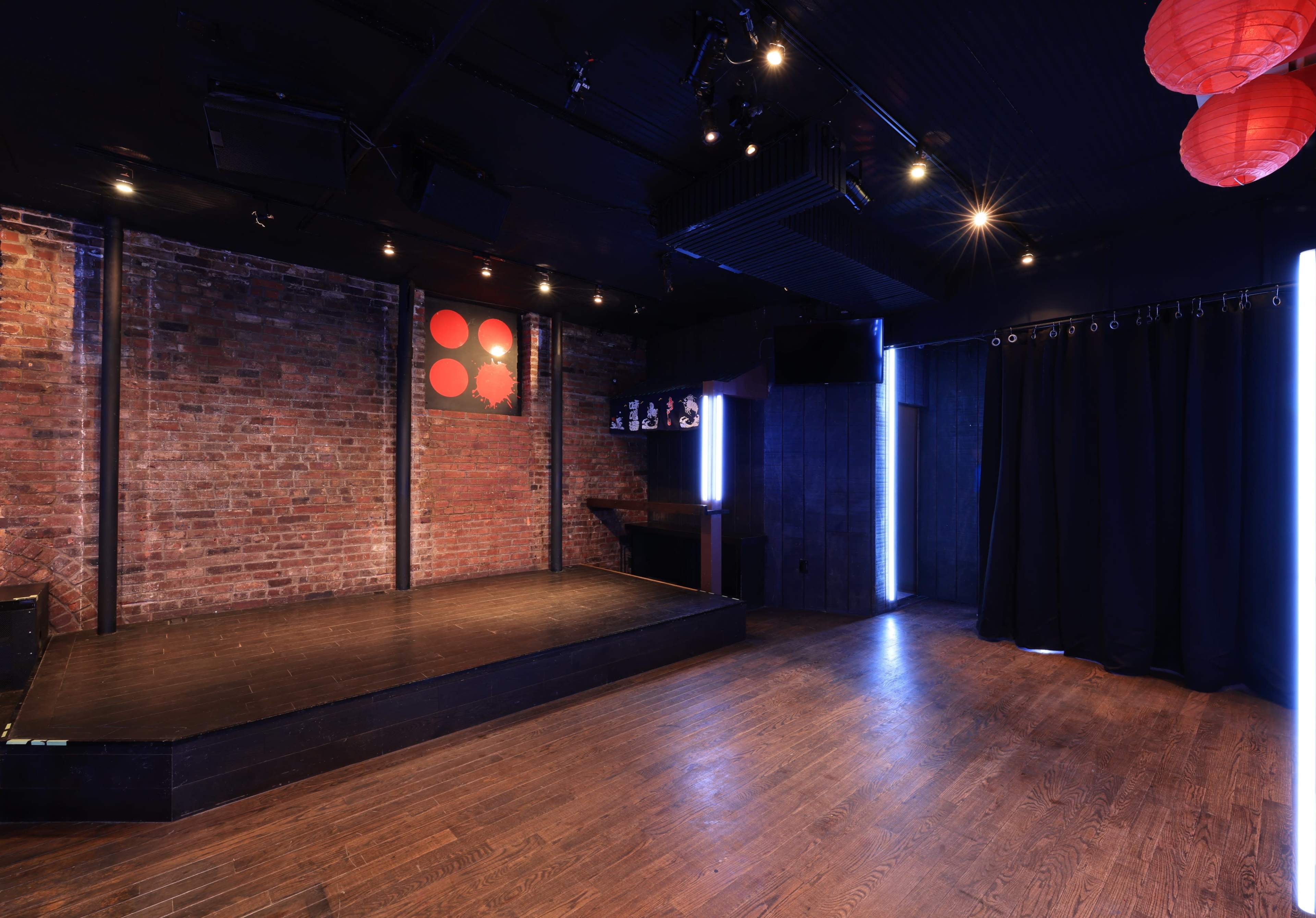 A dimly lit performance space with a wooden stage, exposed brick walls, and red lanterns hanging from the ceiling.
