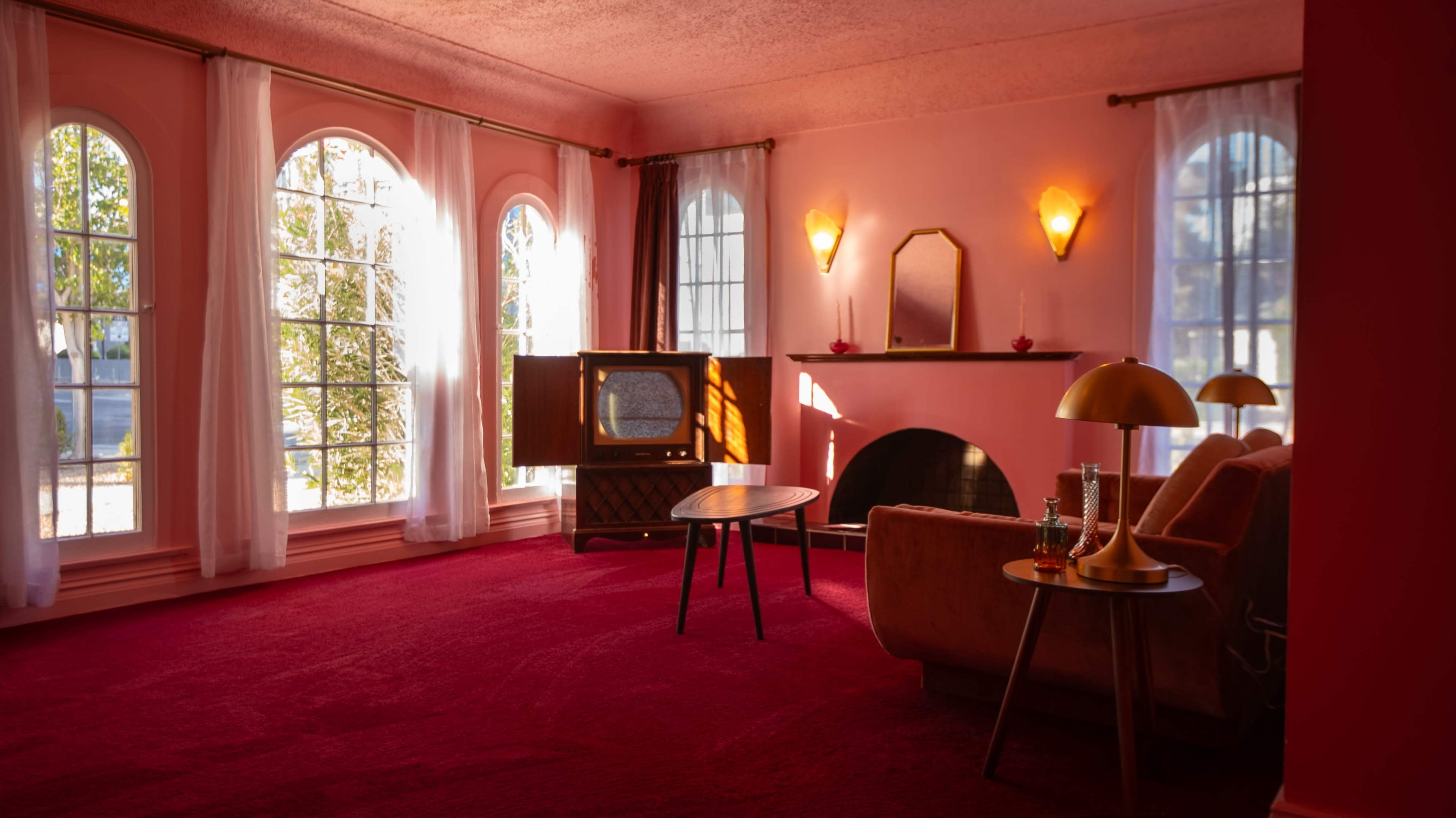 The image shows a cozy living room with large arched windows, pink walls, a vintage television set, a small round table, and a sofa on a red carpet.