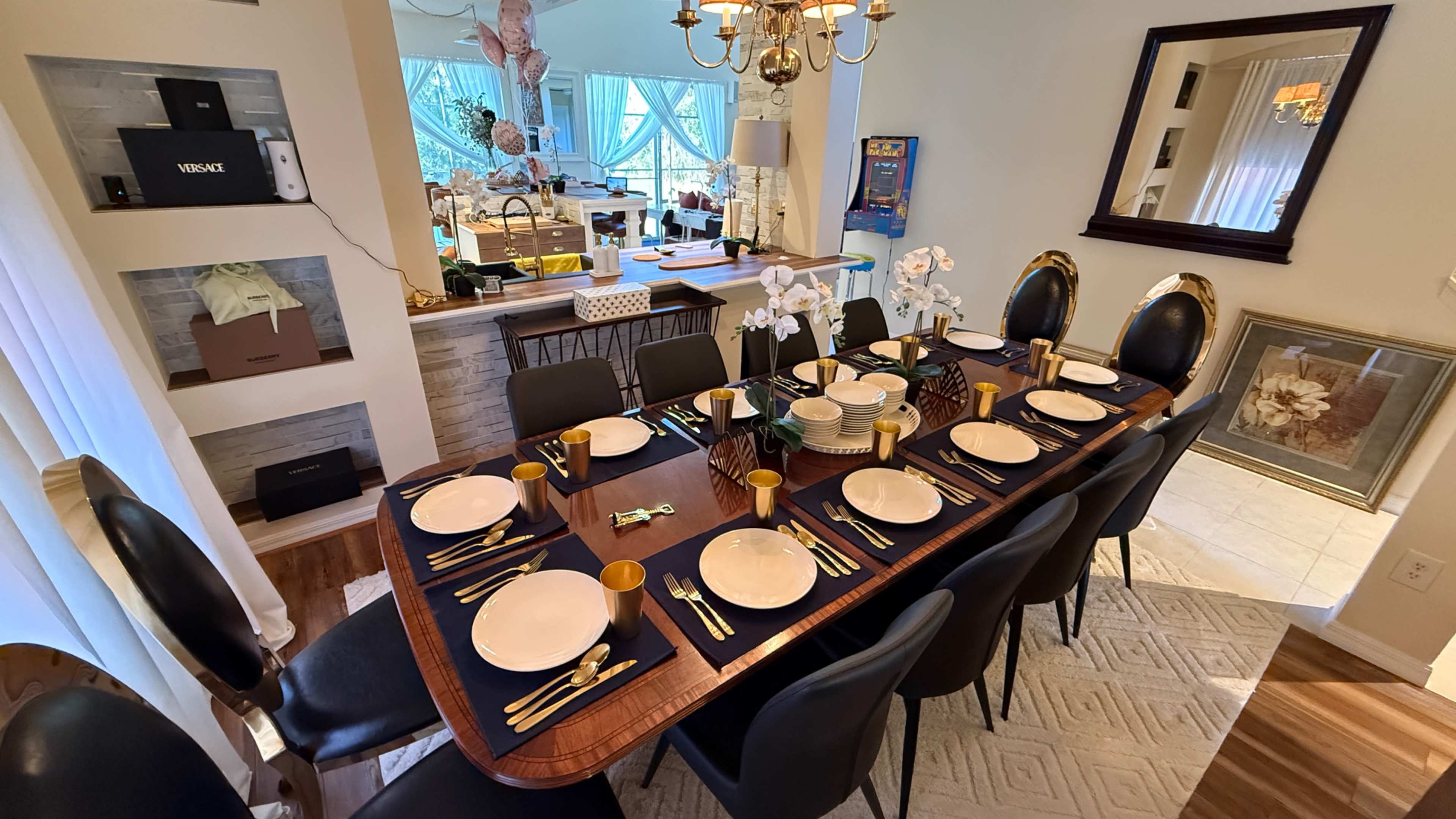 A large dining table is set with white plates and gold utensils, surrounded by black chairs, in a well-lit room with decorative elements and a view of a kitchen area.