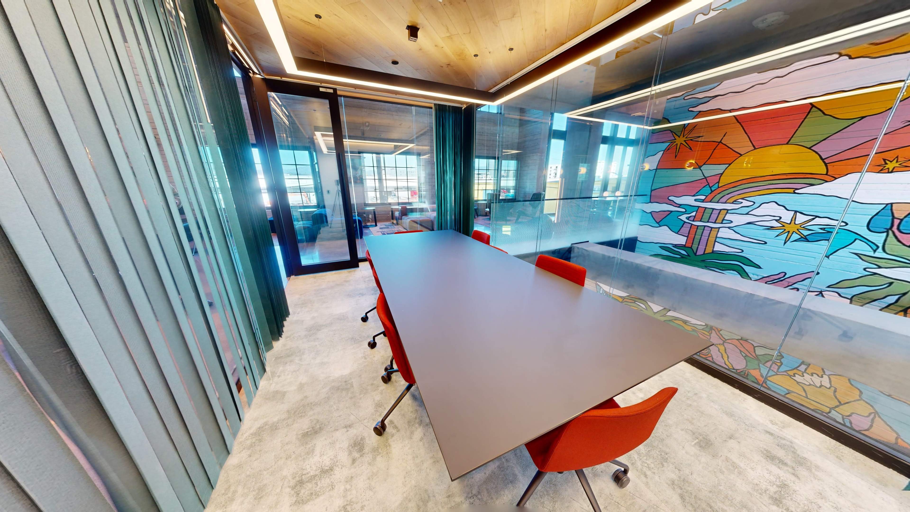 A long conference table with four red chairs is positioned in a glass-walled meeting room featuring a colorful mural on one side.