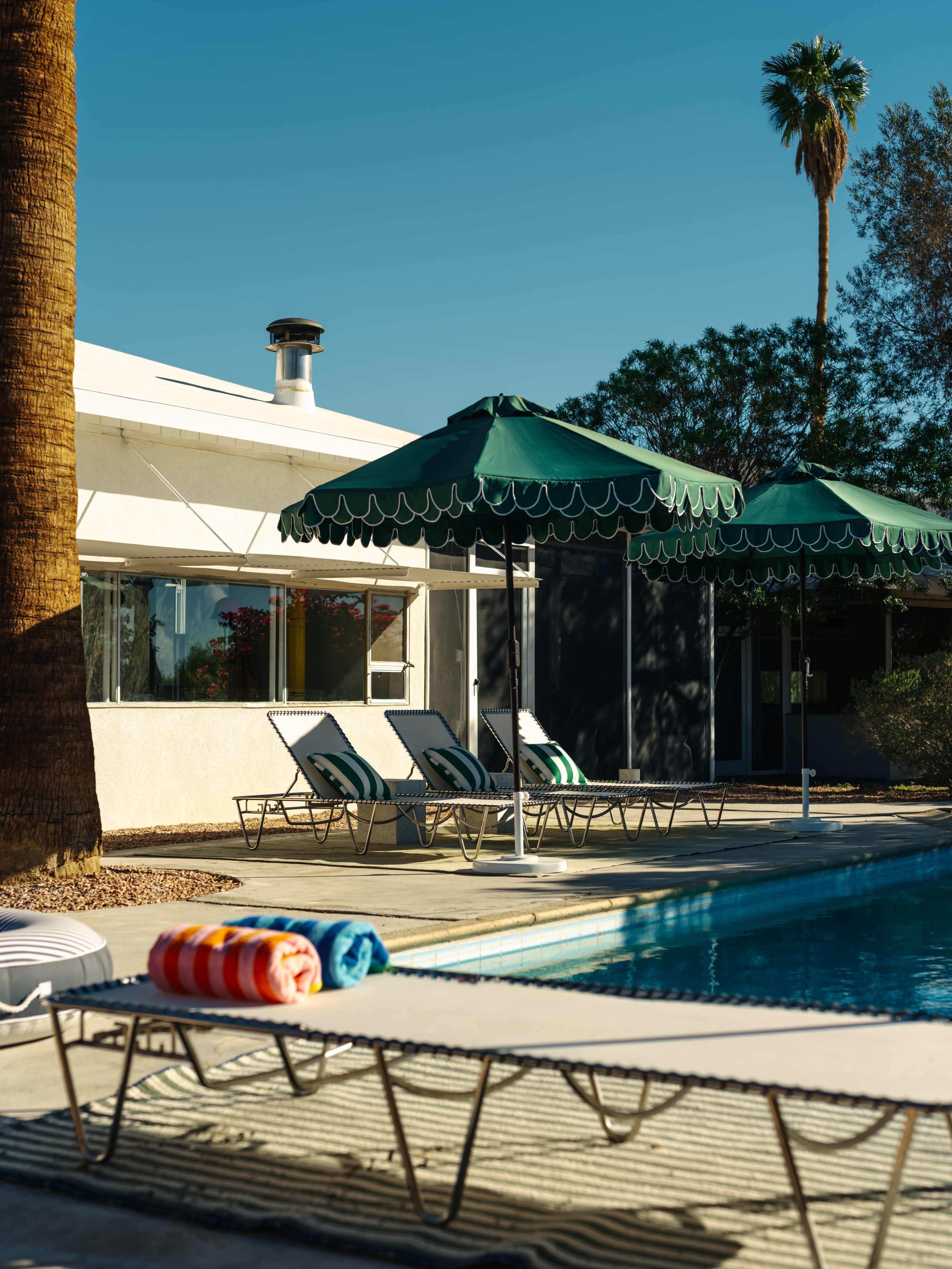 The image shows a pool area with green umbrellas, lounge chairs, and colorful towels beside a clear blue pool under a clear sky.