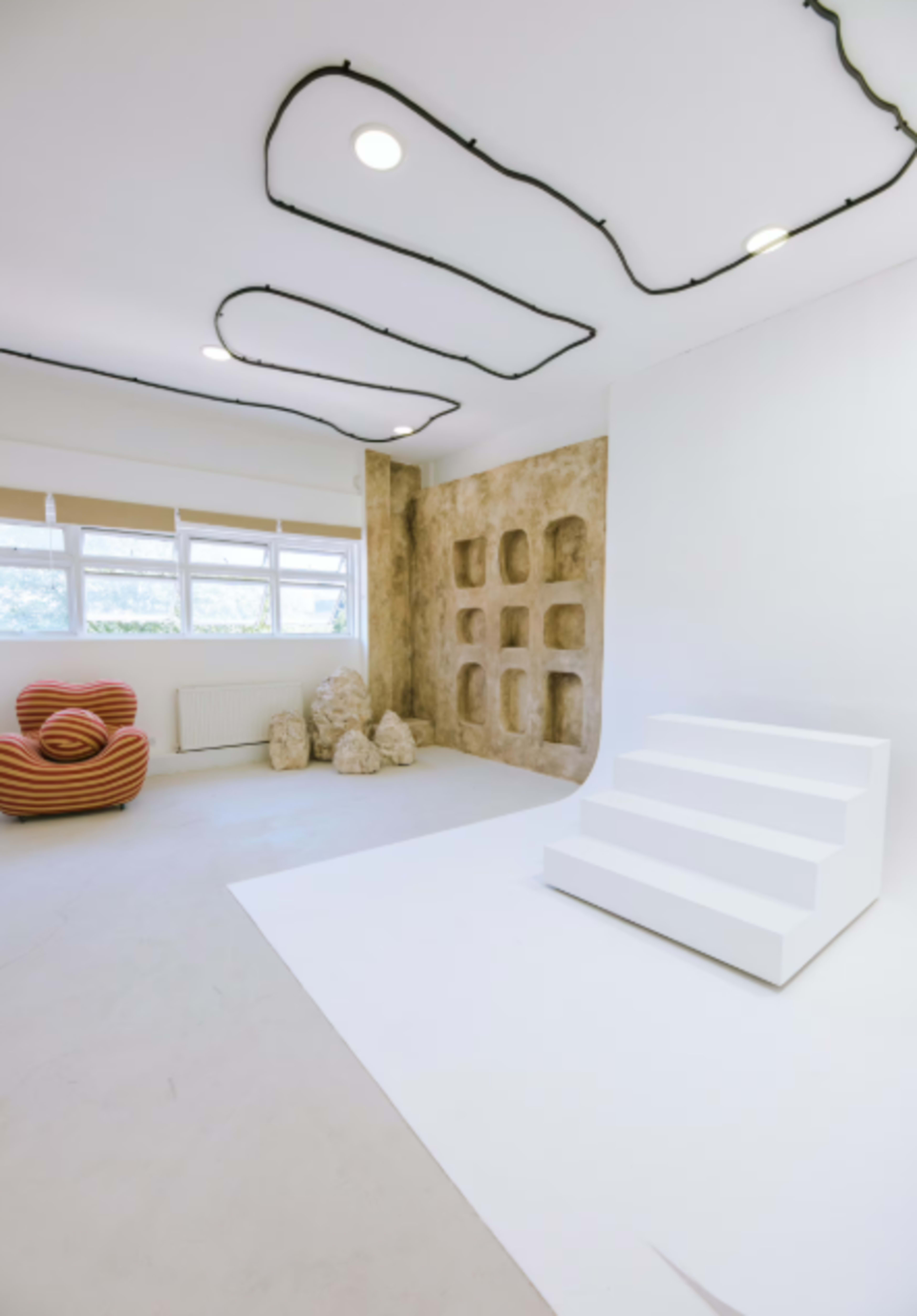 The image shows a spacious, modern room with a textured wall, a set of white stairs, and minimalist furniture including a striped chair.
