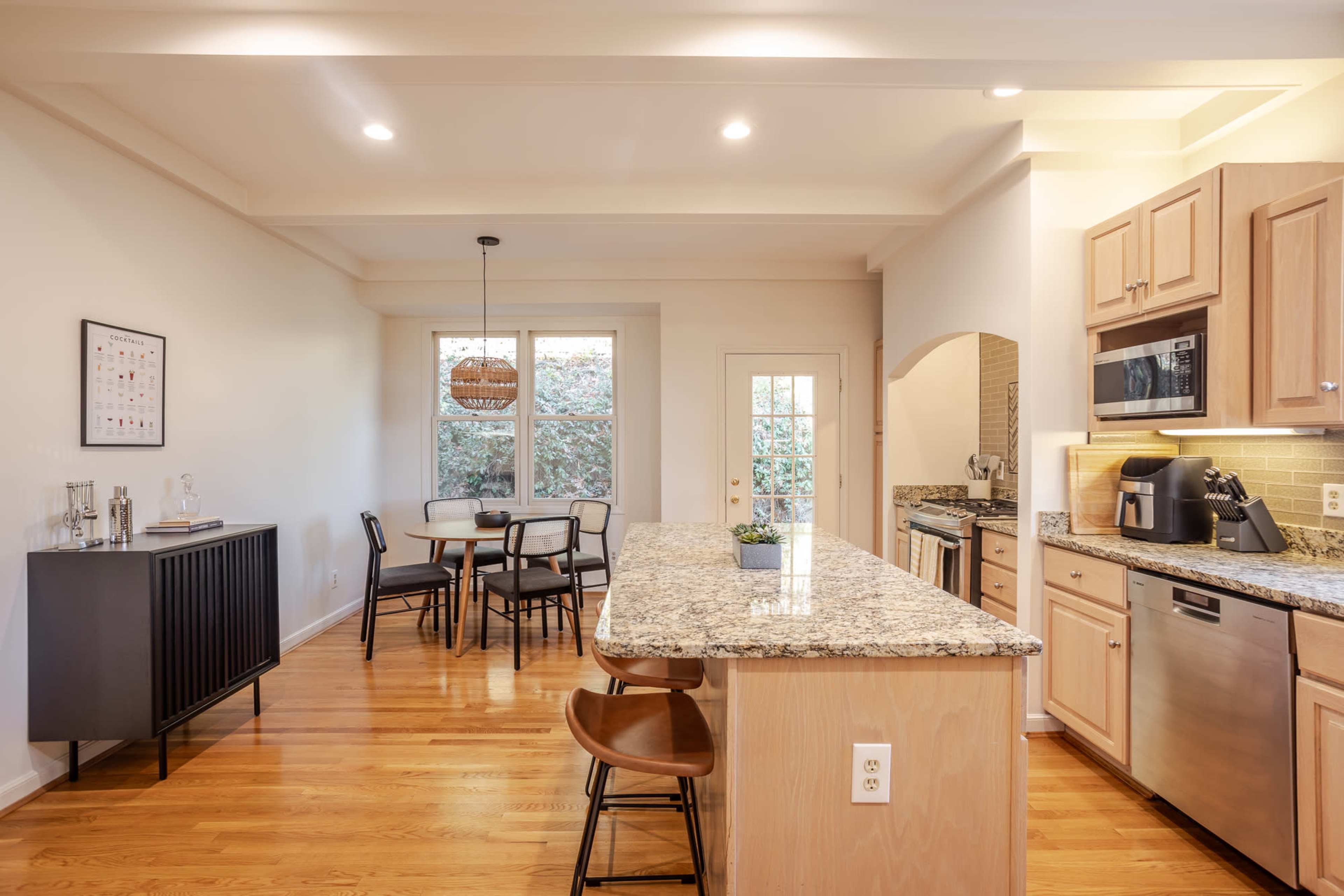 A modern kitchen with wooden cabinets, a granite countertop island, a dining area in the background, and large windows letting in natural light.