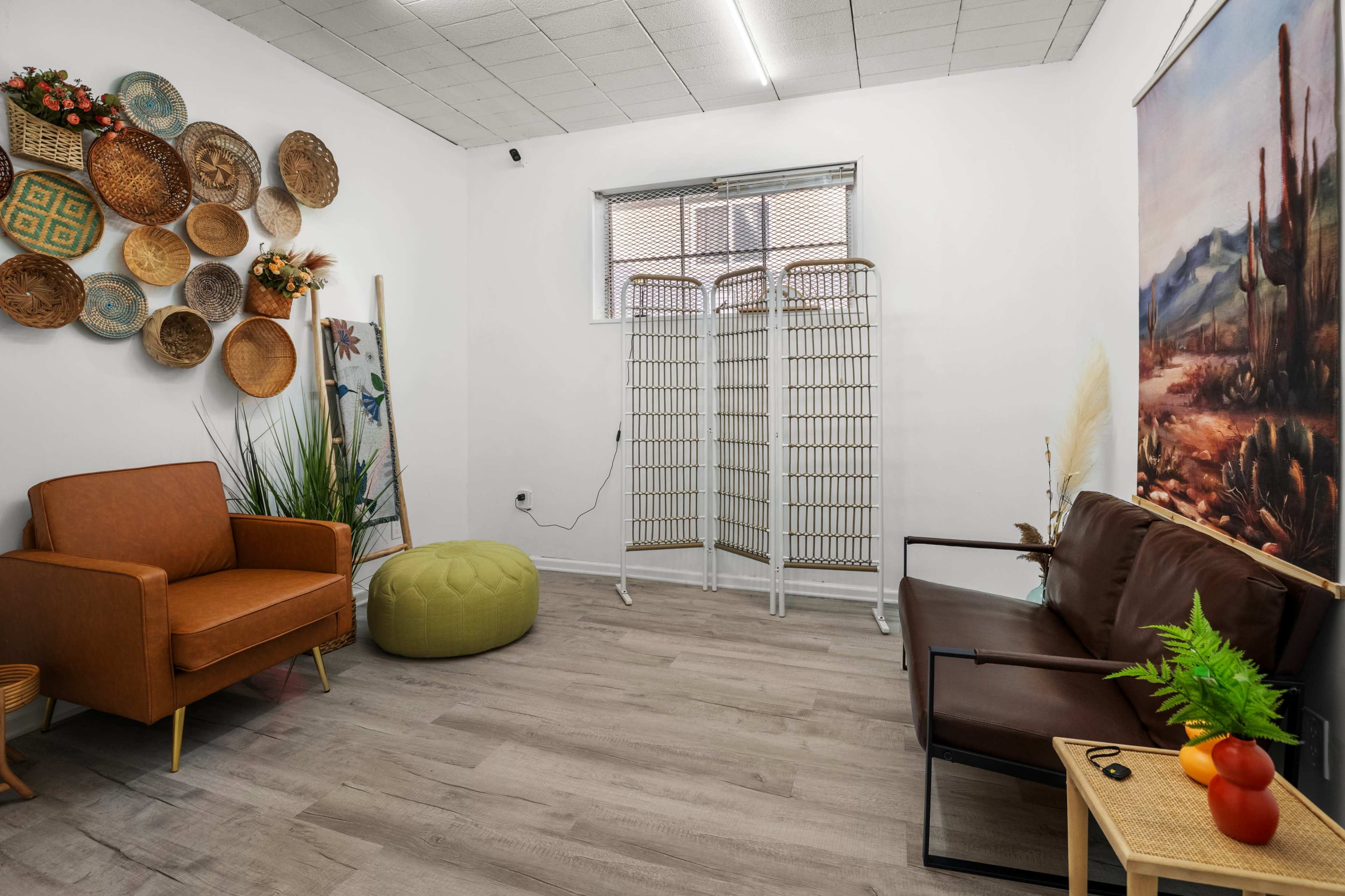 The image shows a small, decorated room featuring a brown couch, an orange armchair, a green pouf, and a wall adorned with woven baskets and a nature-themed painting.