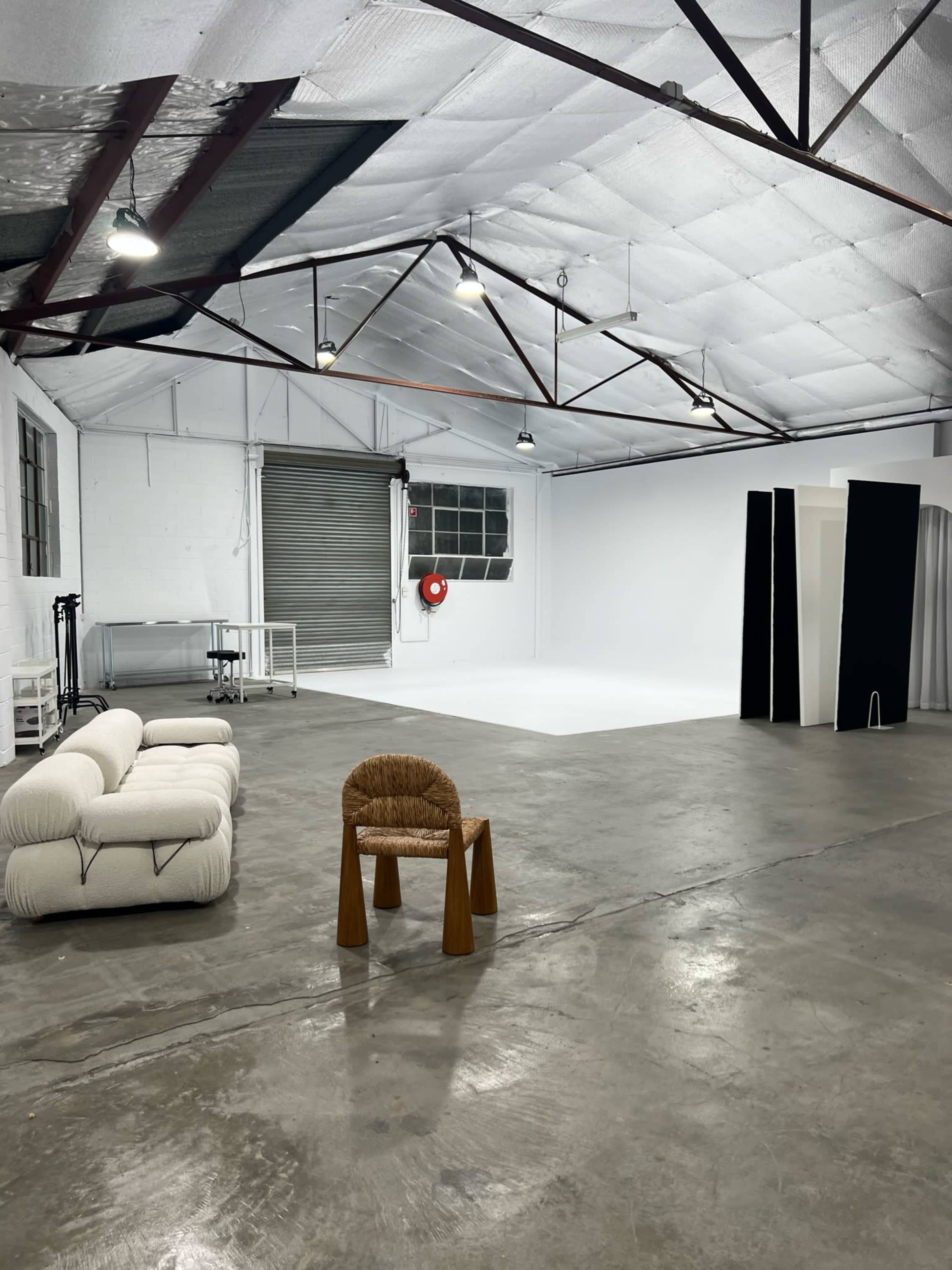 An empty warehouse space with a polished concrete floor, a white backdrop, and minimal furniture including a chair and a couch.