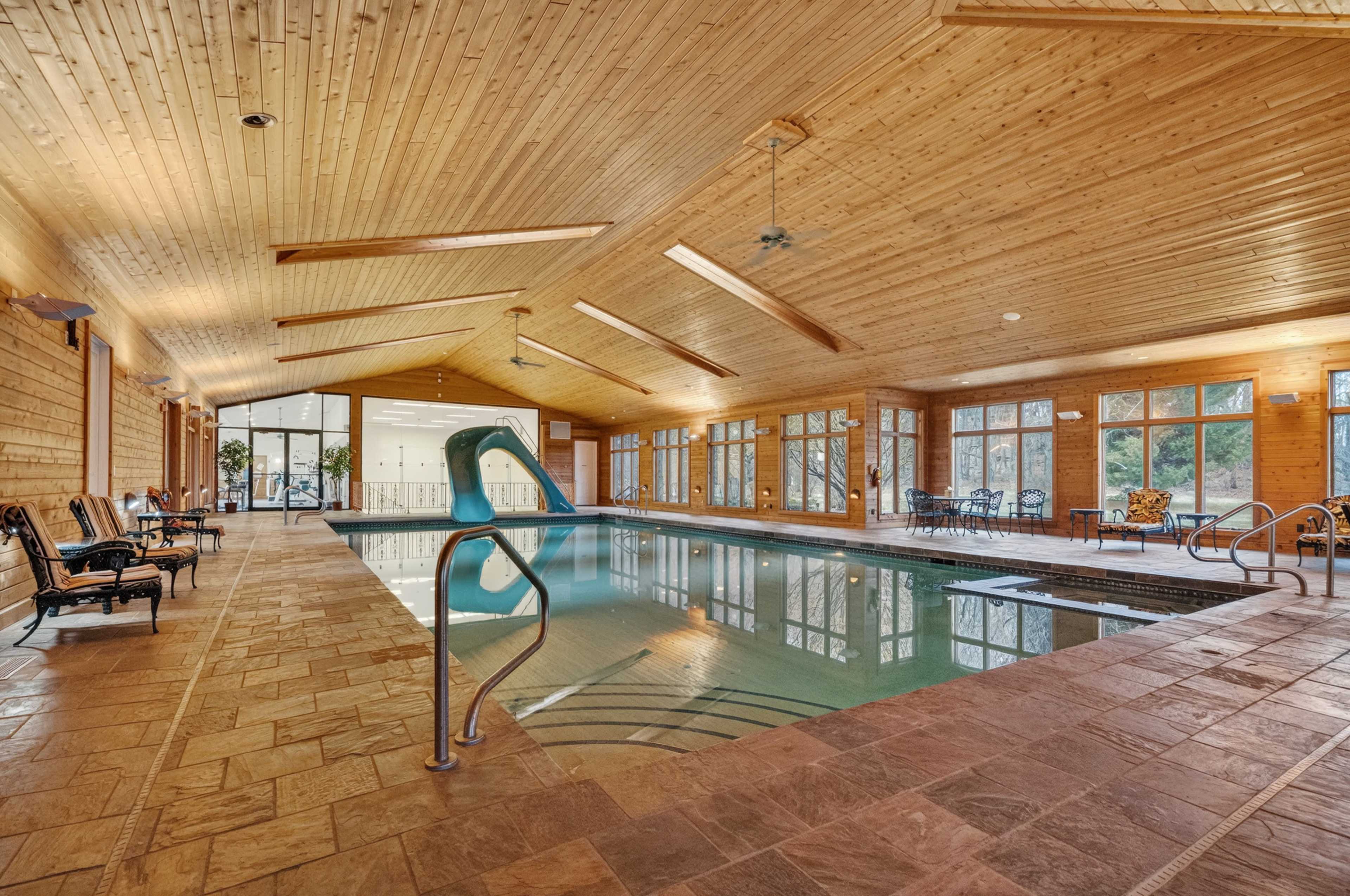The image shows an indoor swimming pool area with a wooden ceiling, several lounge chairs, and large windows allowing natural light to enter.