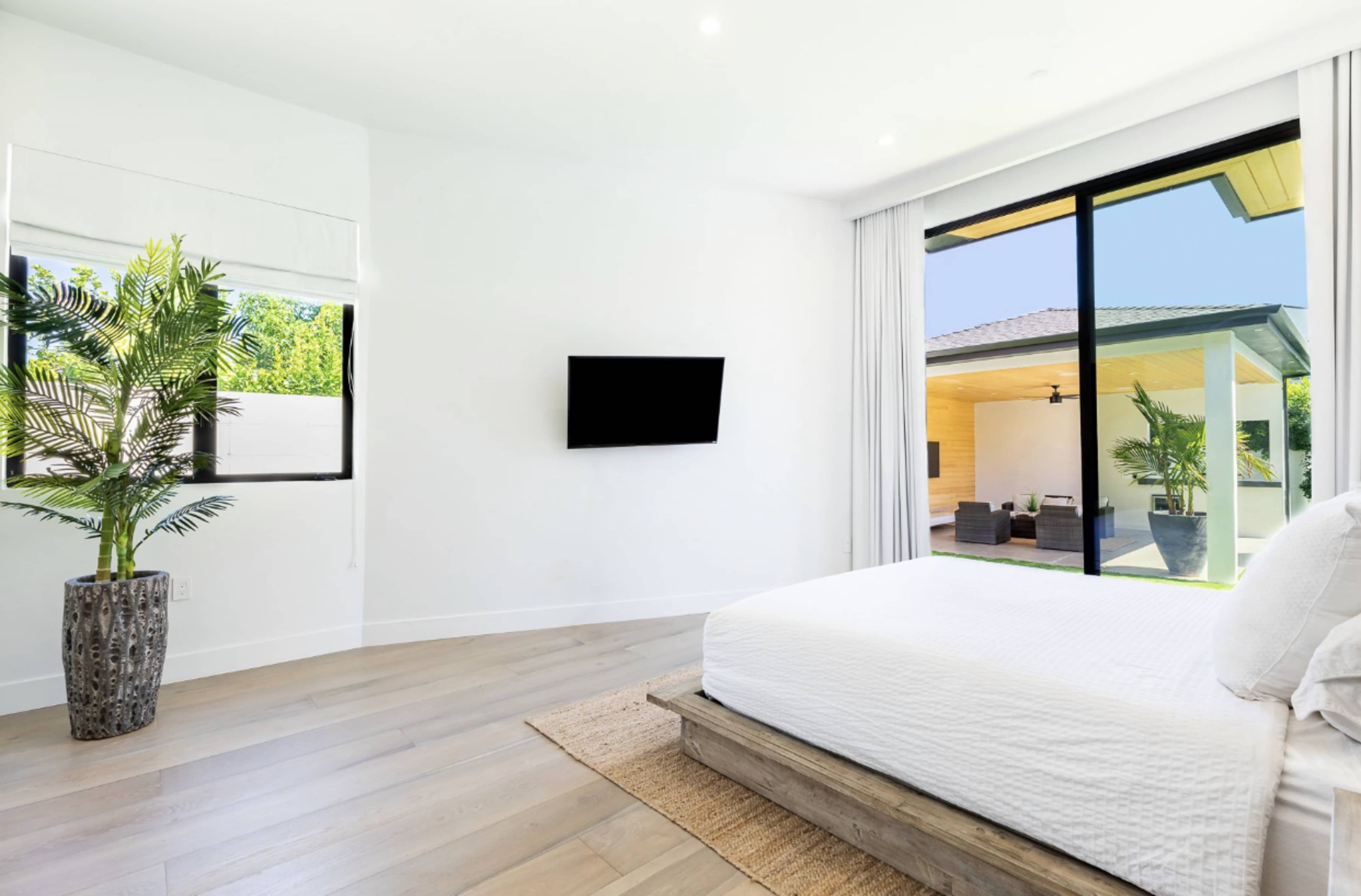 A modern bedroom features large windows with a view of an outdoor seating area, a flat-screen TV mounted on the wall, and a bed with a wooden frame.