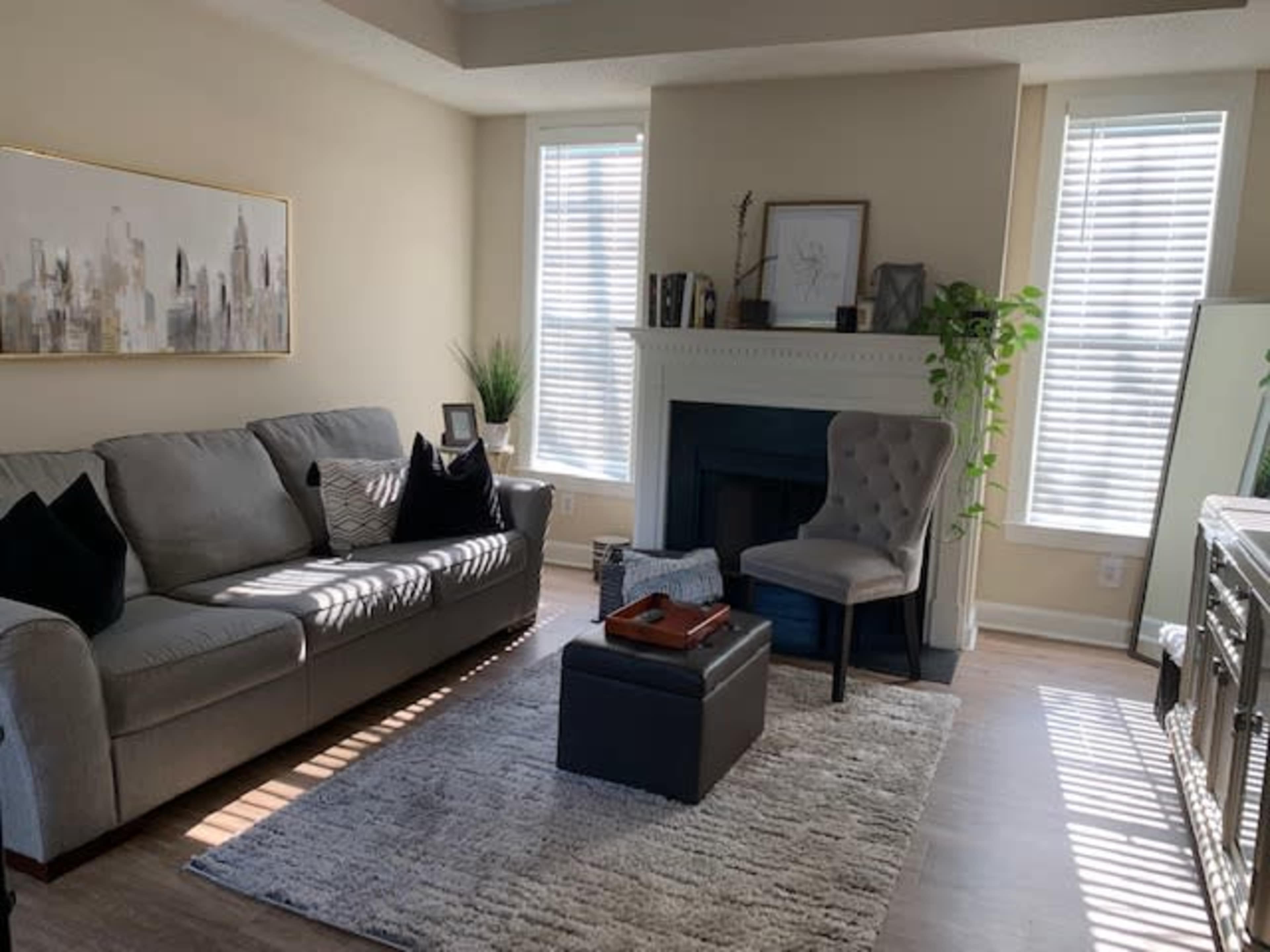 A living room features a gray sofa, a light-colored rug, a small table, and a chair beside a fireplace, with natural light streaming through the window blinds.