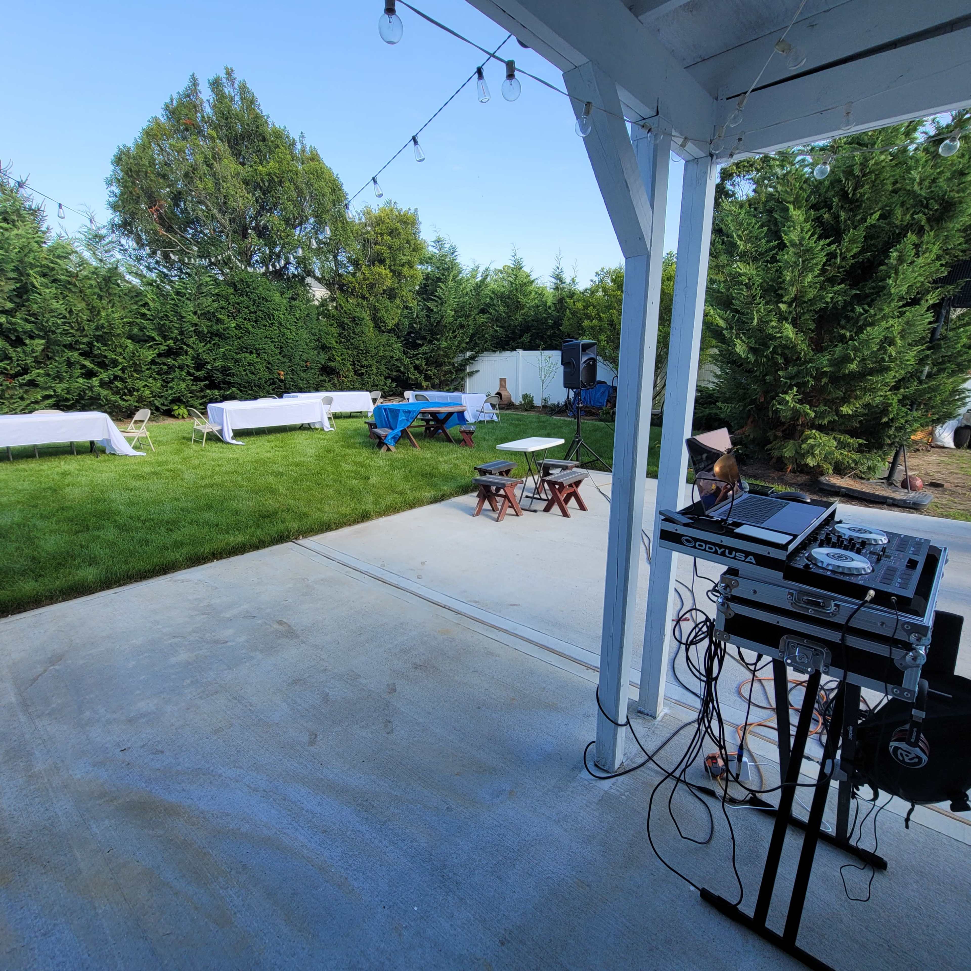 An outdoor party setup features tables covered in white and blue tablecloths, a DJ station with equipment, and string lights overhead in a grassy backyard.