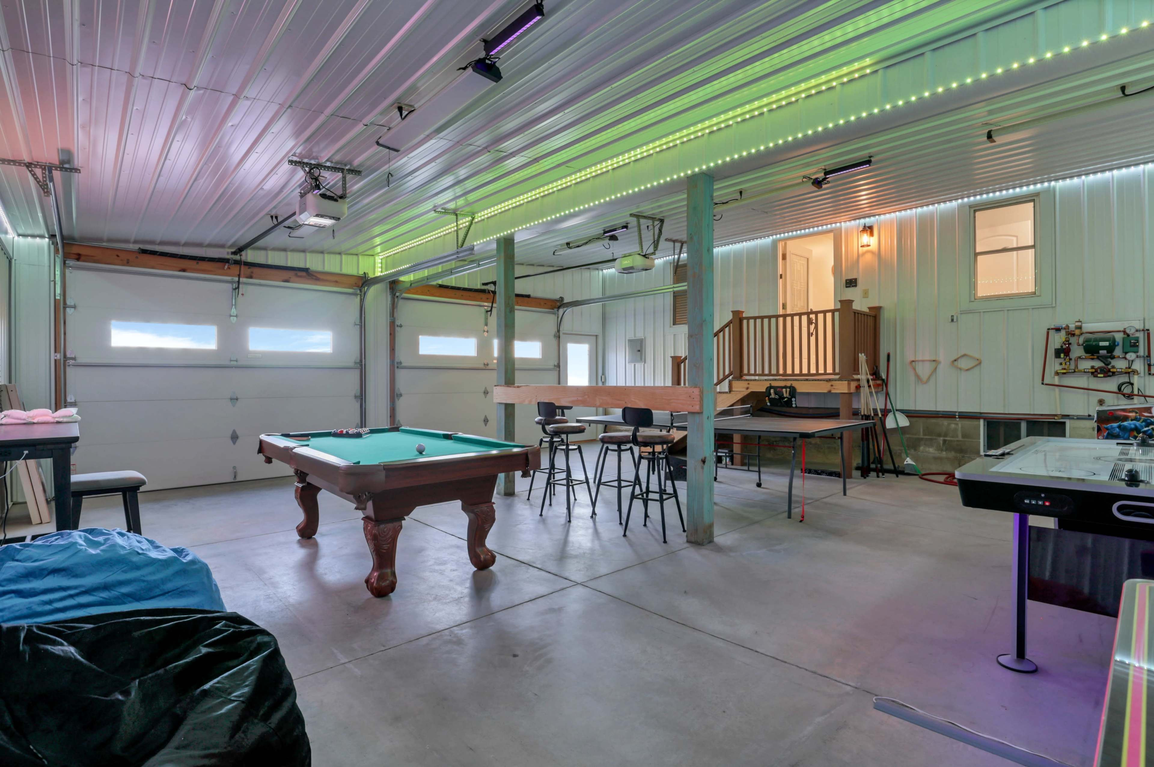 A spacious garage that features a billiards table, bar-style seating, and various recreational equipment, illuminated by multicolored LED lights along the ceiling.