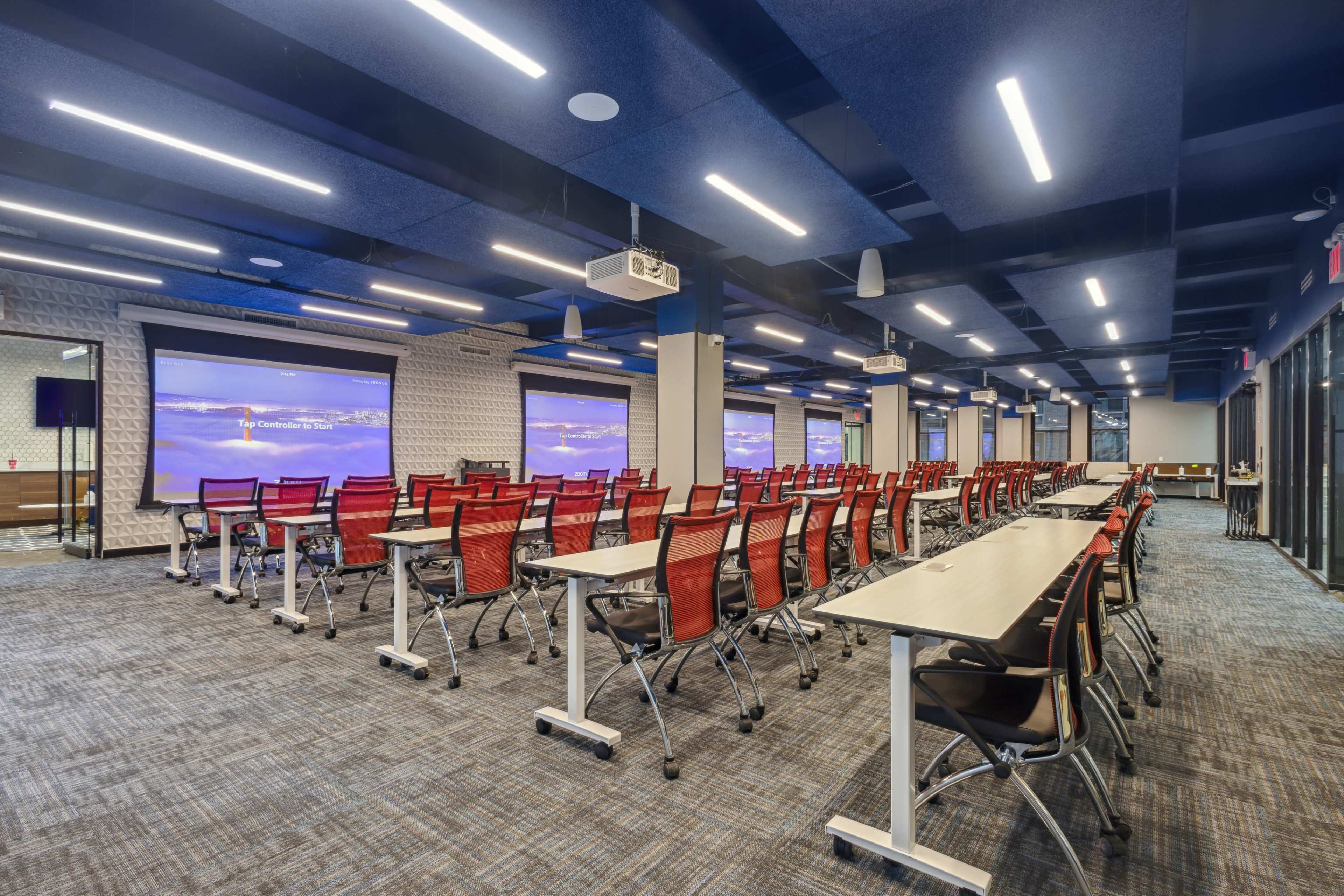 The image shows a modern conference room with rows of tables and red chairs, featuring large screens at the front for presentations.