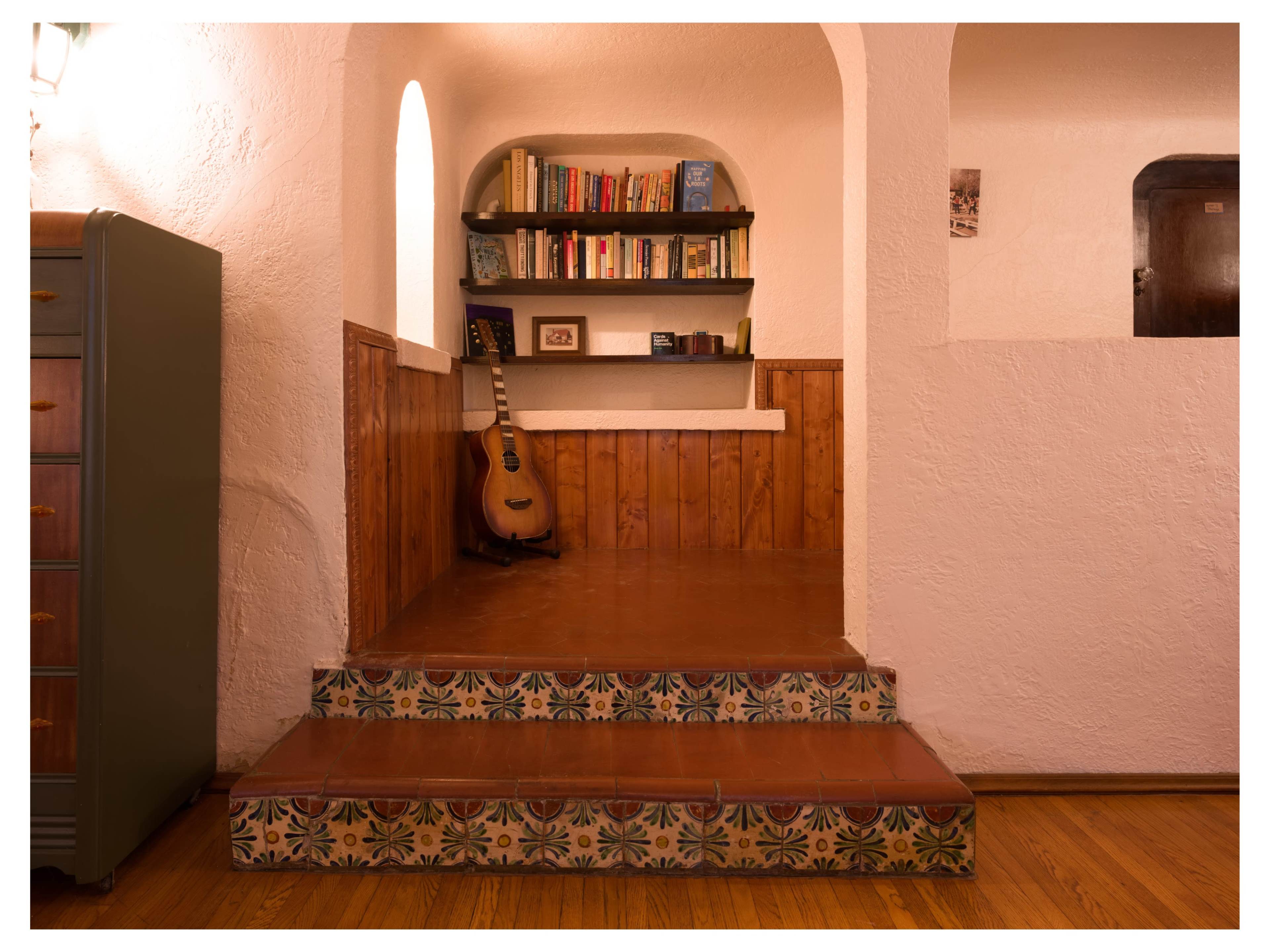 The image depicts a stairway leading up to a small alcove with a bookshelf and a guitar against a textured white wall.
