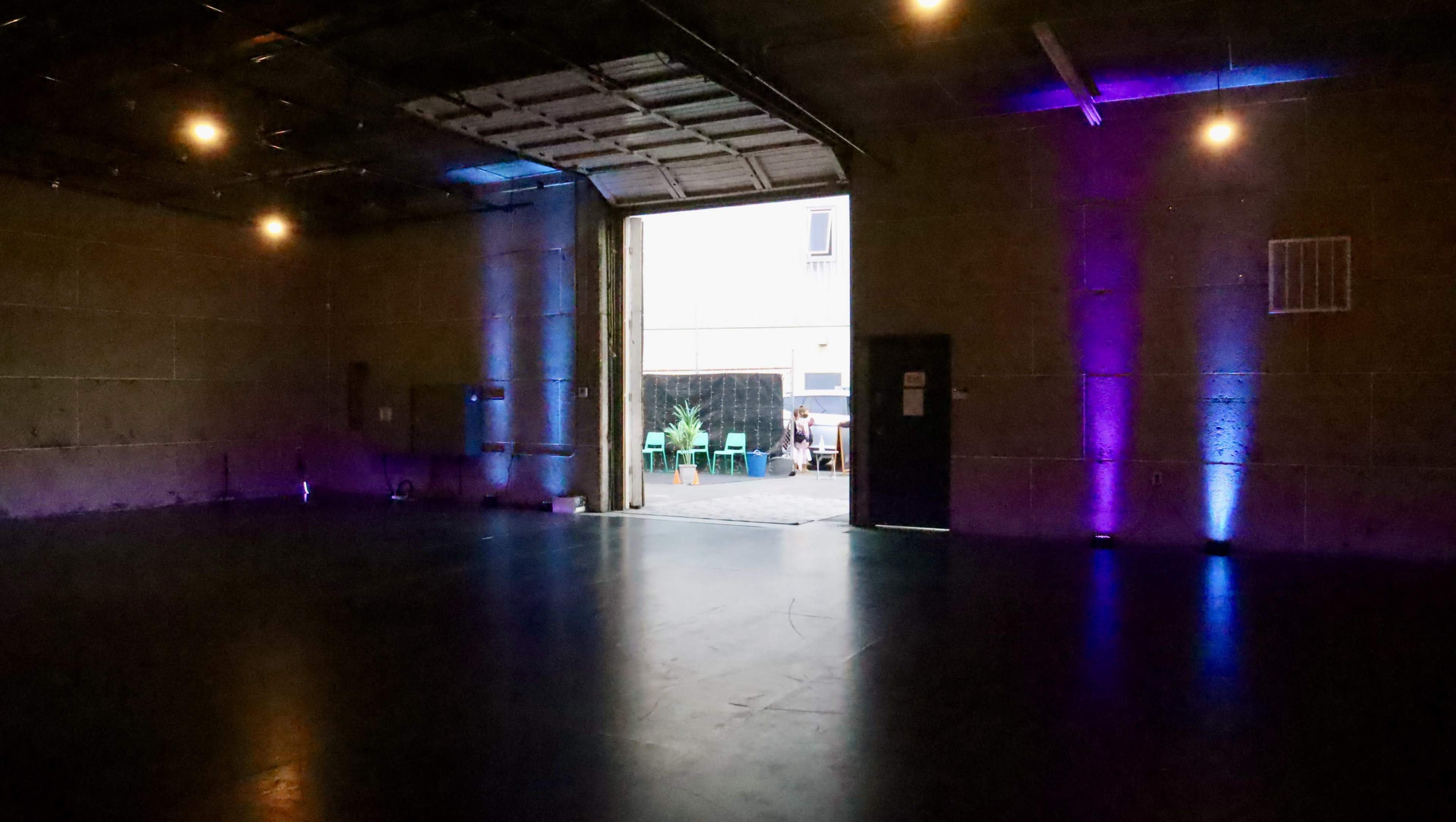 An empty industrial space is illuminated by colored lights, with an open door leading to a courtyard featuring green chairs and plants.