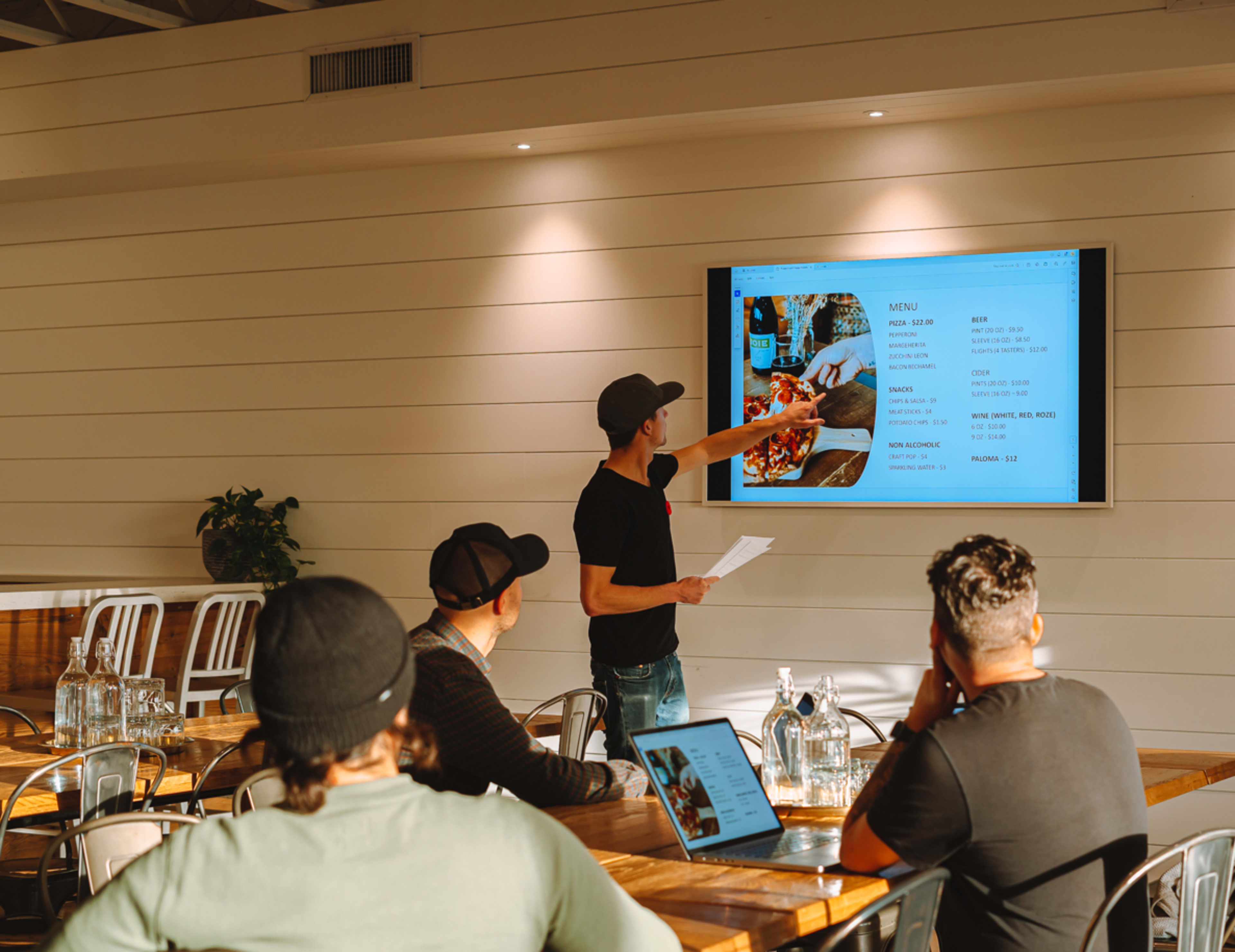 A man points at a presentation screen displaying a menu while three colleagues watch in a bright meeting room.