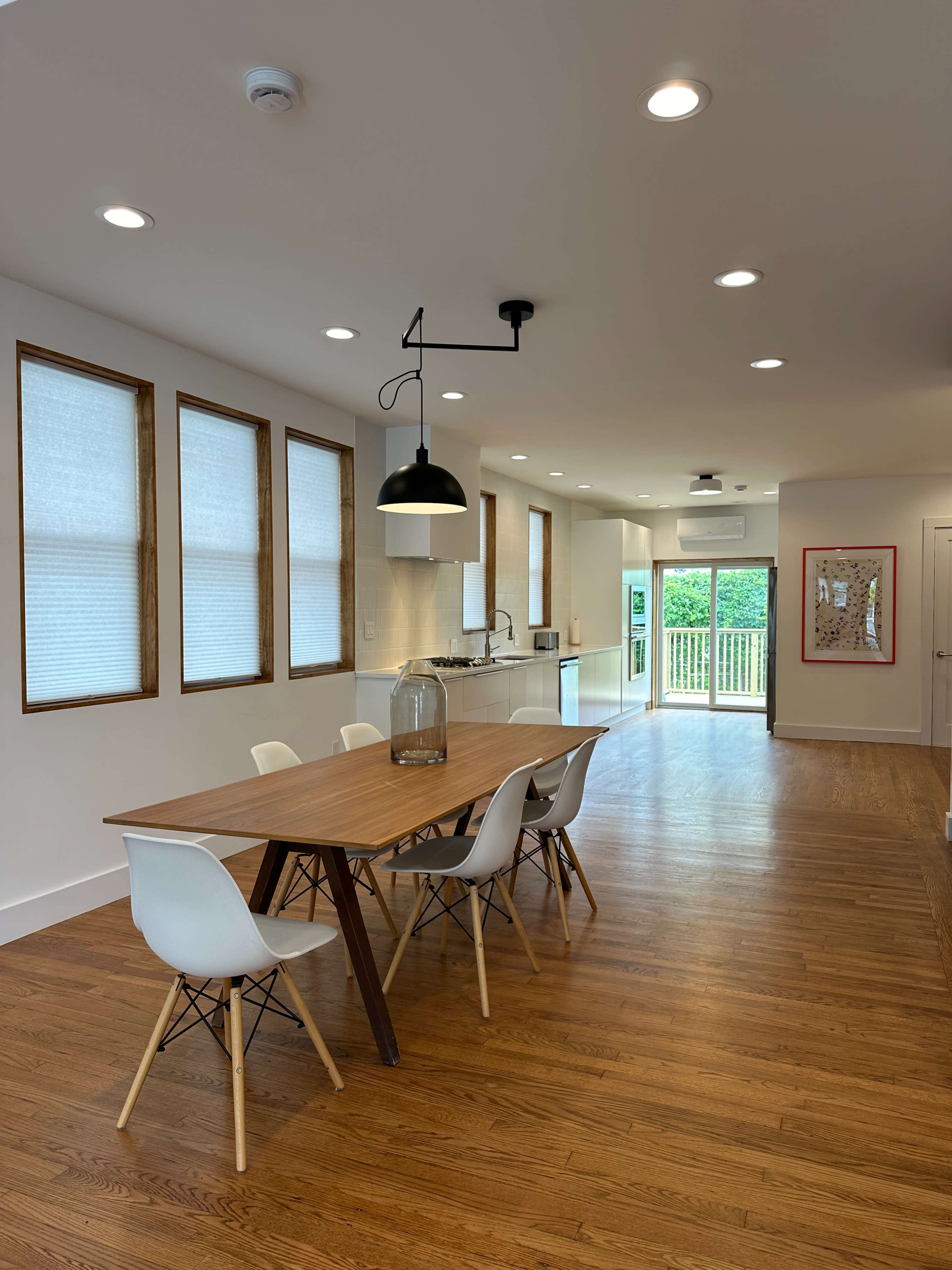 A dining area with a wooden table and white chairs is situated in a brightly lit room featuring large windows, leading to an outdoor deck.