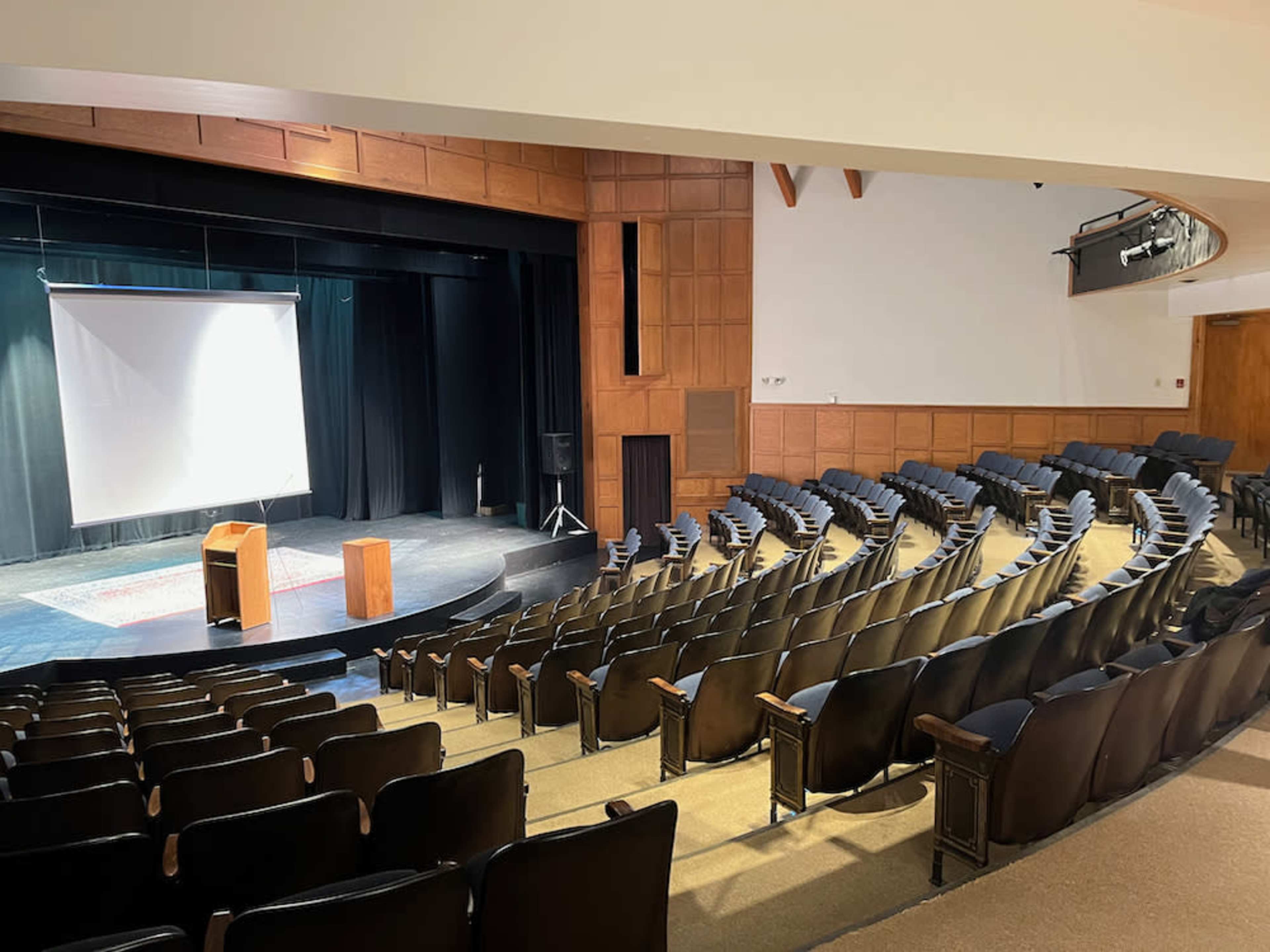 An empty theater with rows of black seats facing a stage equipped with a projection screen and a wooden podium.