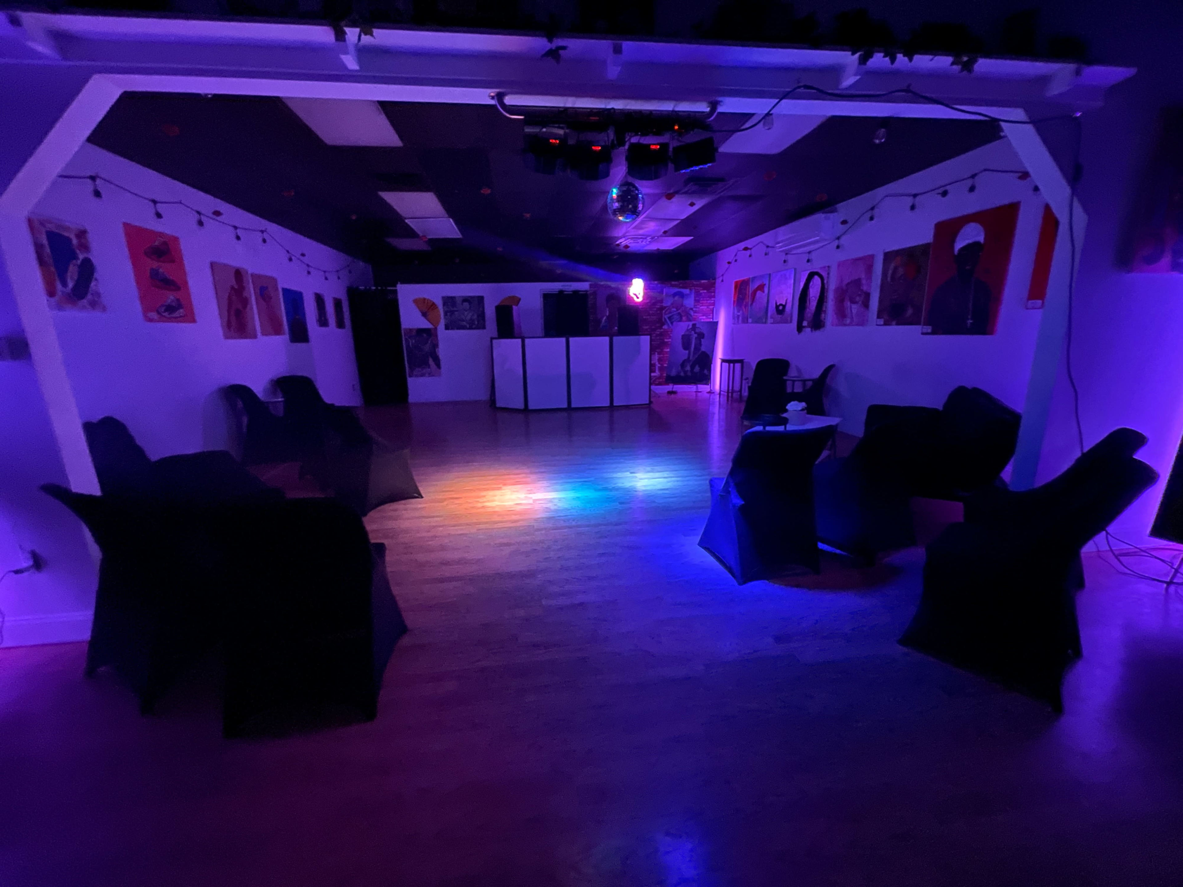 The image shows an empty room with dark walls, colorful lighting, and several chairs positioned around a central area.