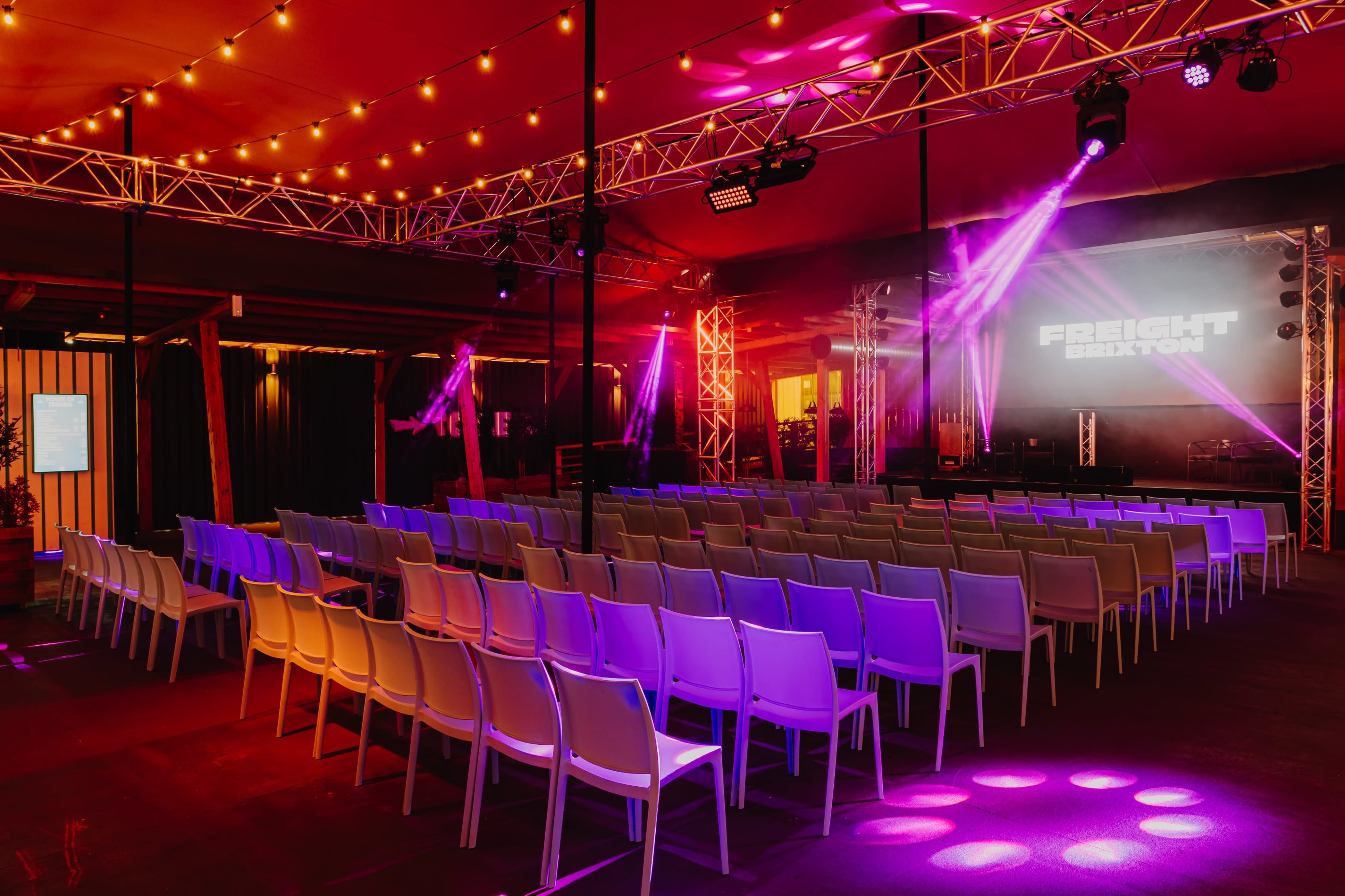 The image shows a concert venue set up with rows of white chairs facing a stage, illuminated by colorful lights and a large screen displaying "FREIGHT."
