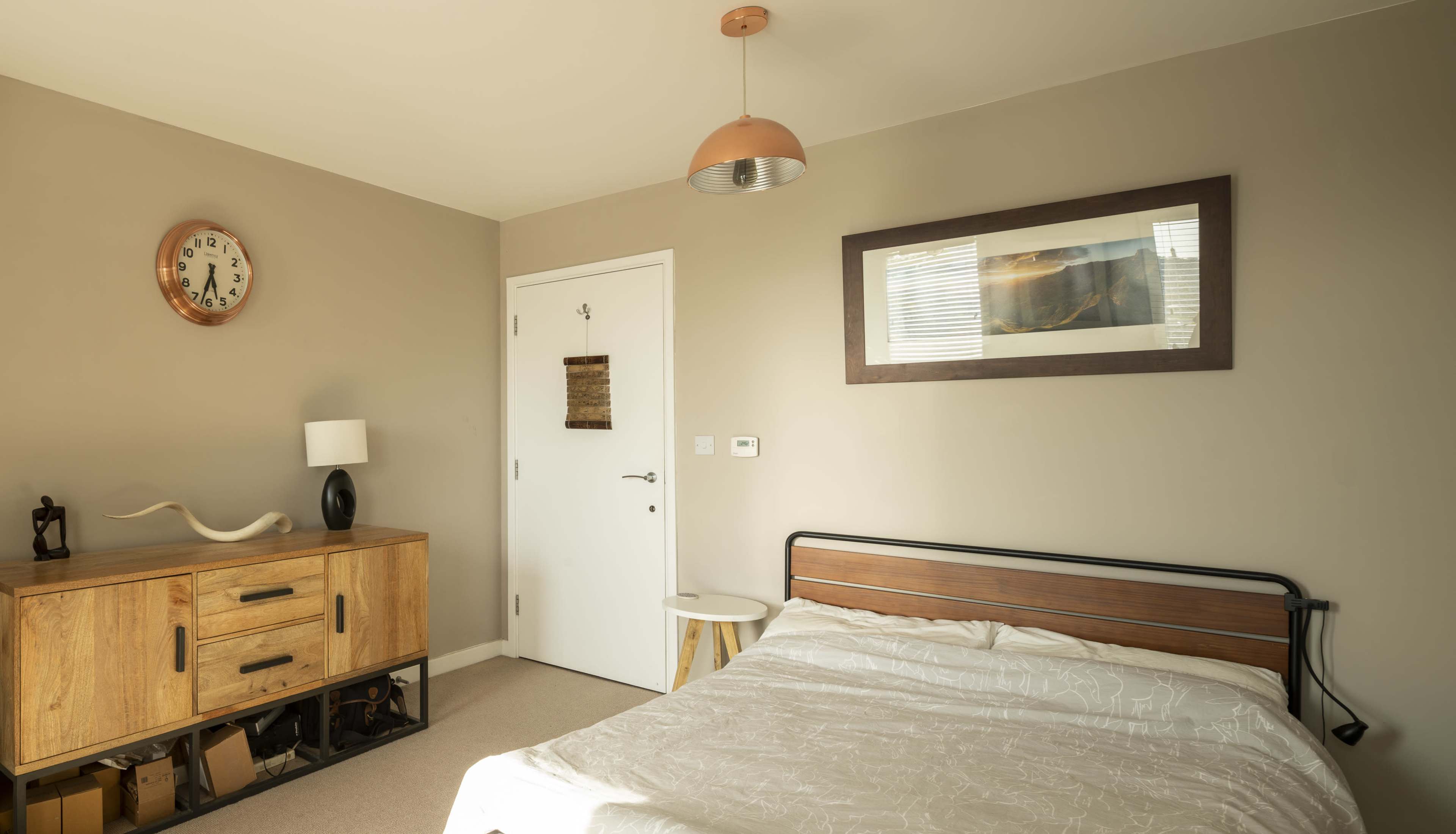 The image shows a neatly arranged bedroom featuring a wooden dresser, a bedside table, and a double bed with a light-colored bedspread against a neutral wall.