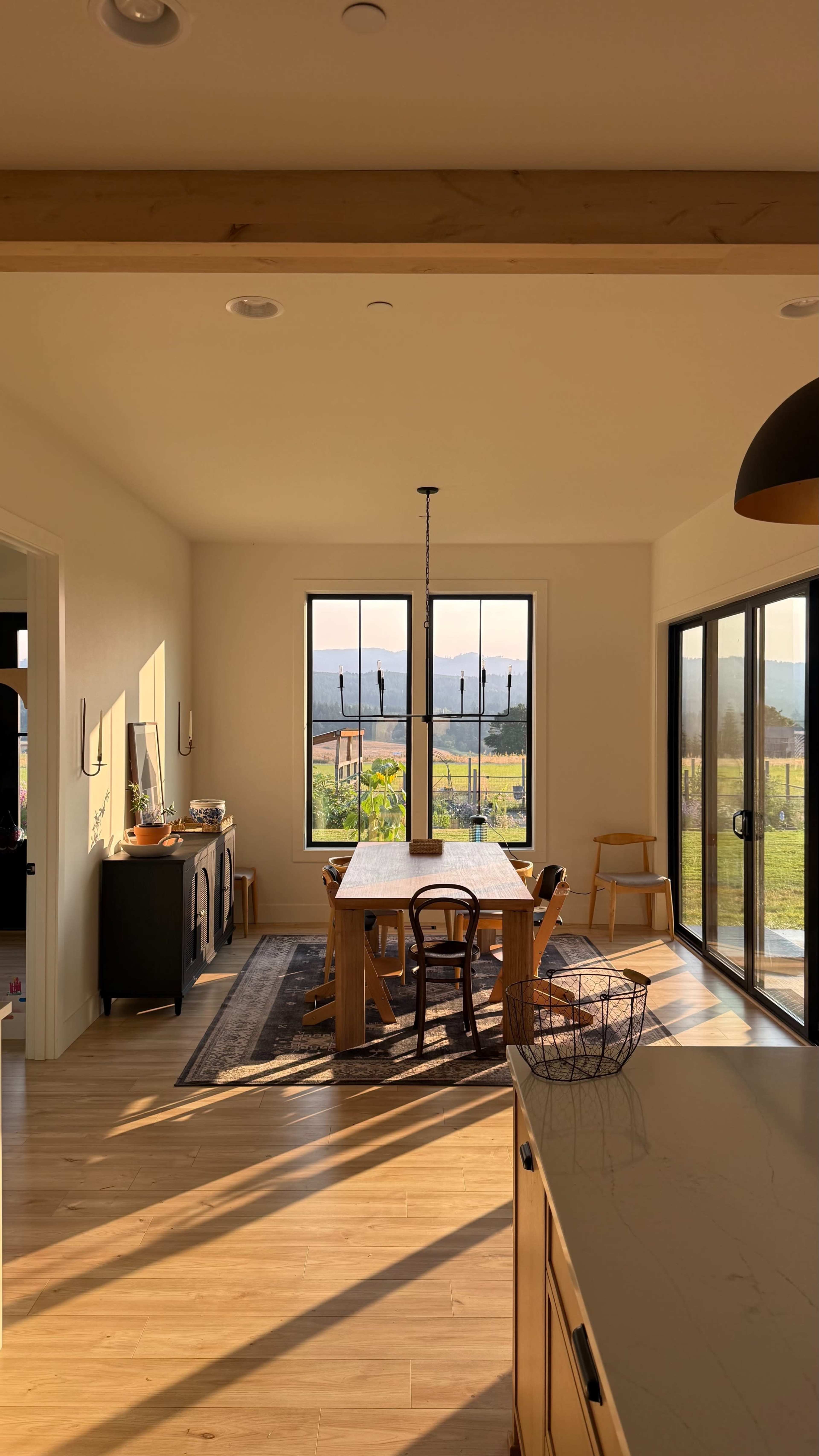A dining area features a long wooden table, chairs, and large windows overlooking a scenic landscape.