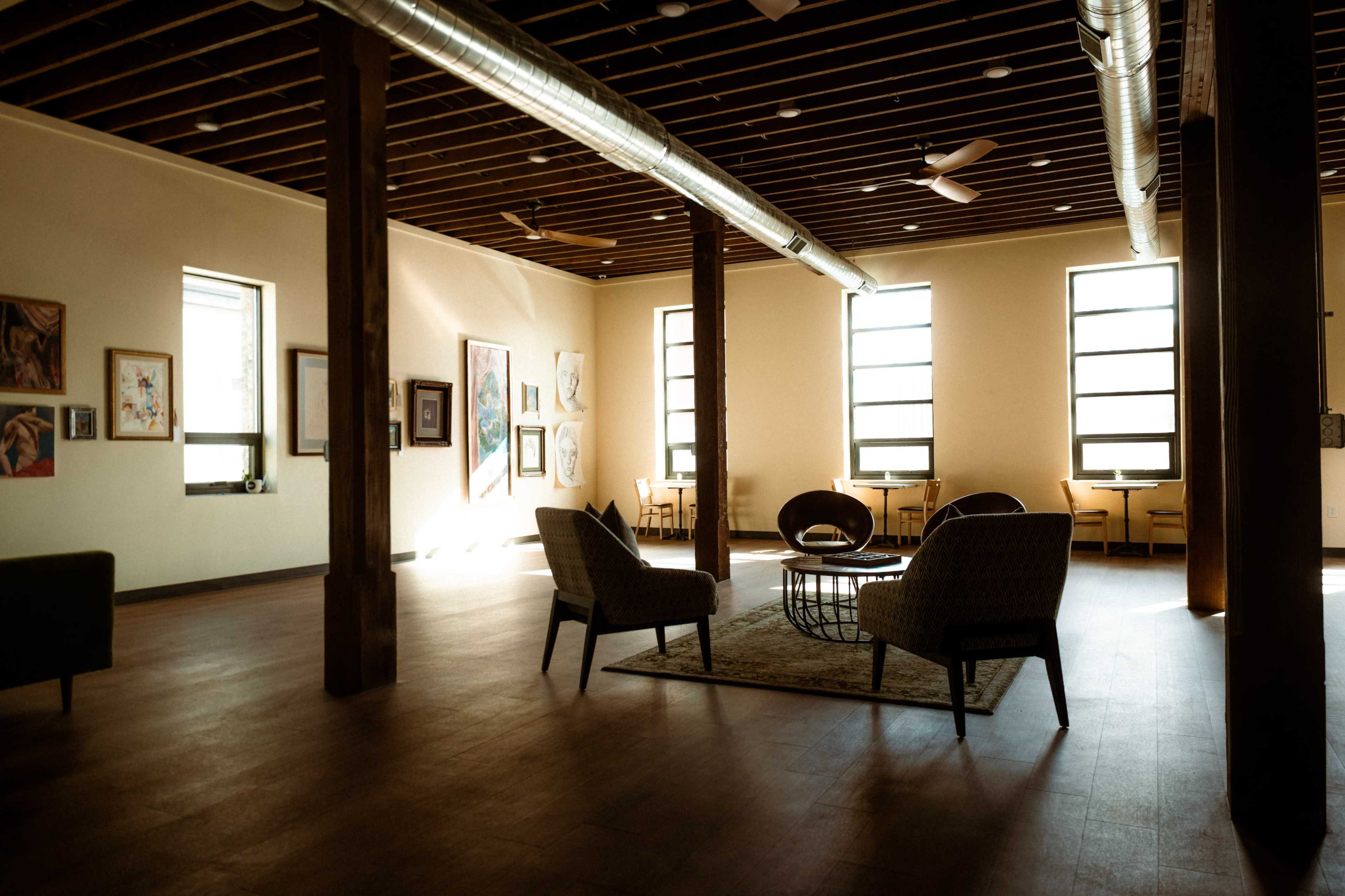 The image shows a spacious room with wooden columns, large windows, and furniture arranged around a central rug.
