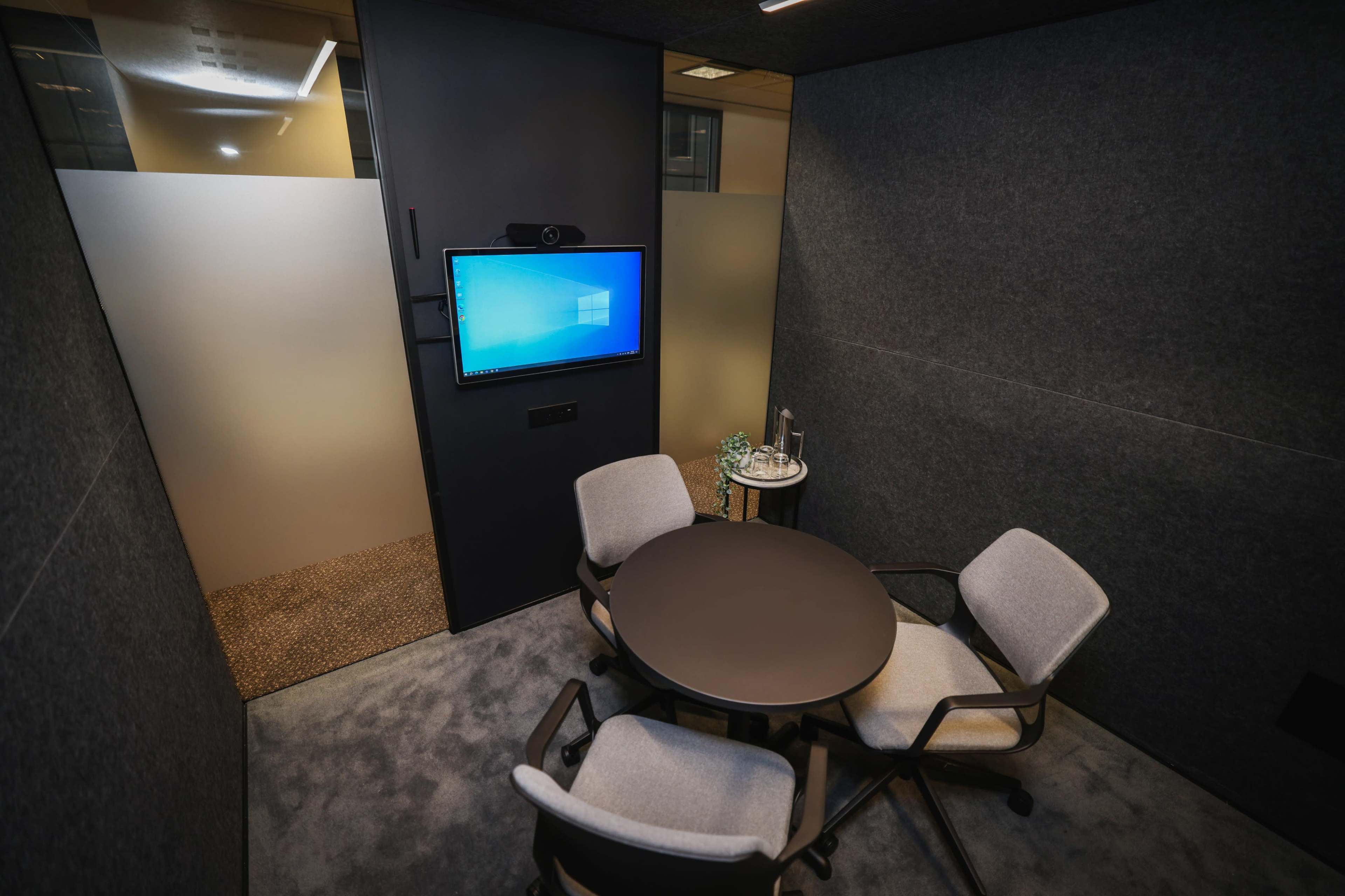 The image shows a small meeting room with a round table, three chairs, a monitor mounted on the wall, and a glass door.