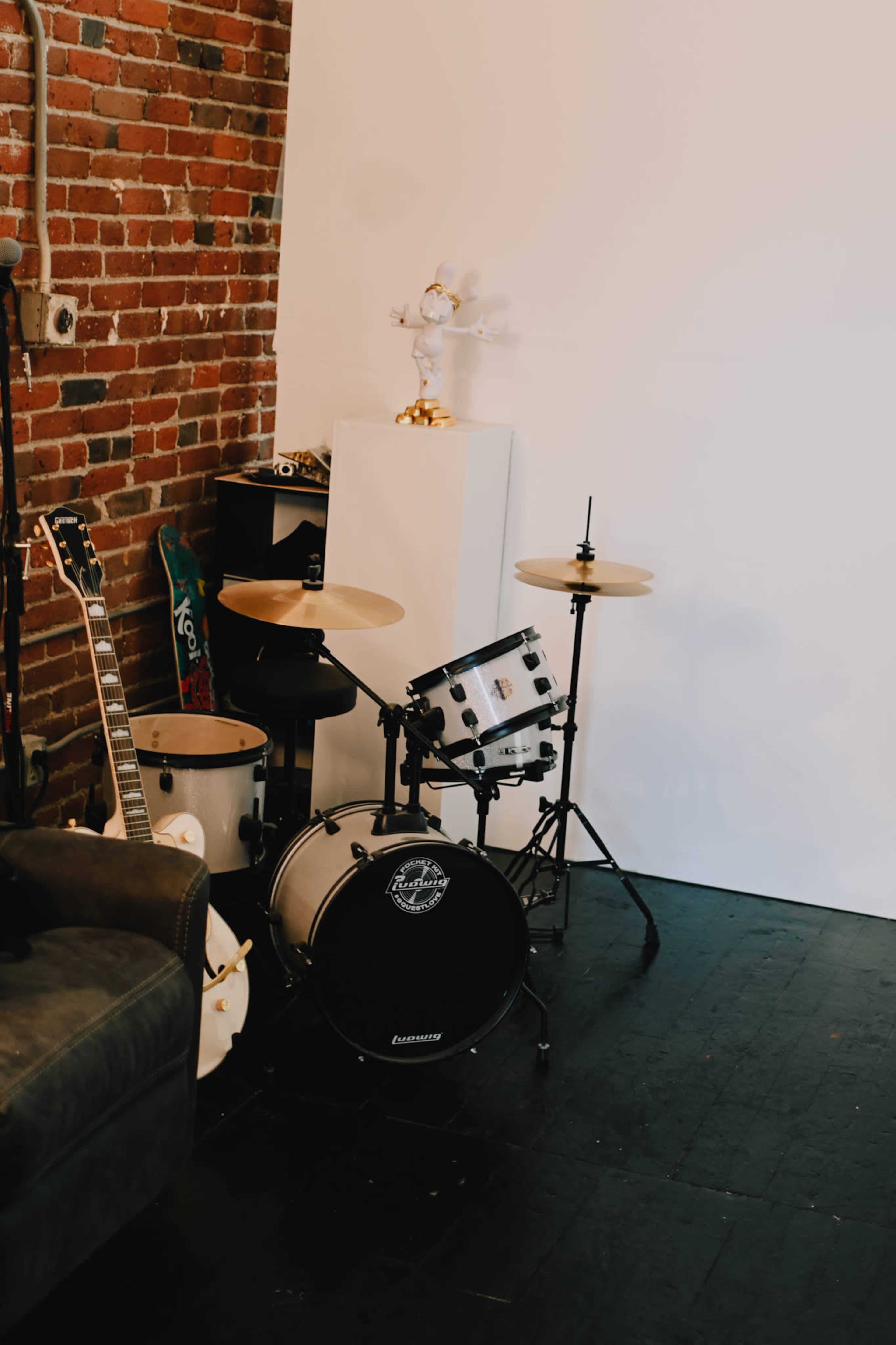 A corner of a room featuring a drum set, a guitar, and a small white pedestal displaying a decorative item.