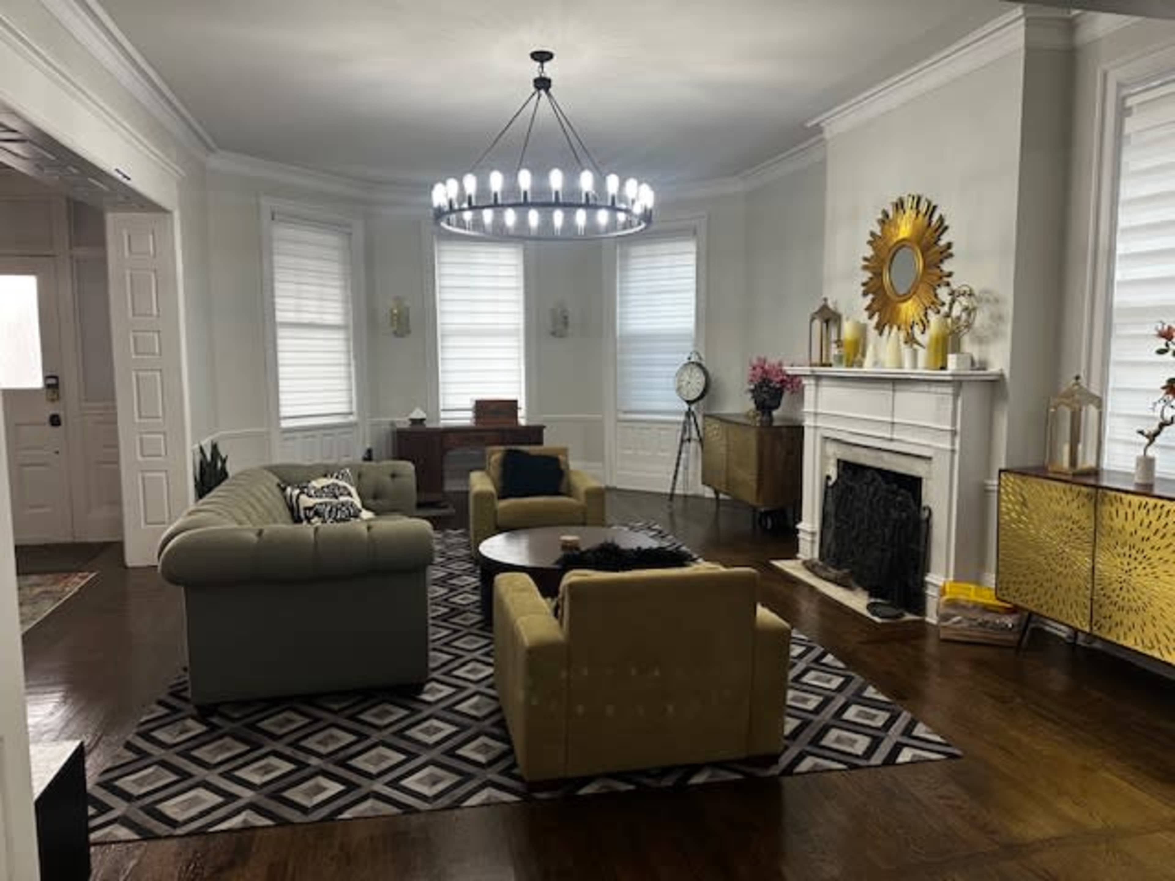 The living room features a central chandelier, a circular coffee table, two armchairs, and a sofa, all arranged around a patterned rug, with a fireplace and decorative elements on the walls.