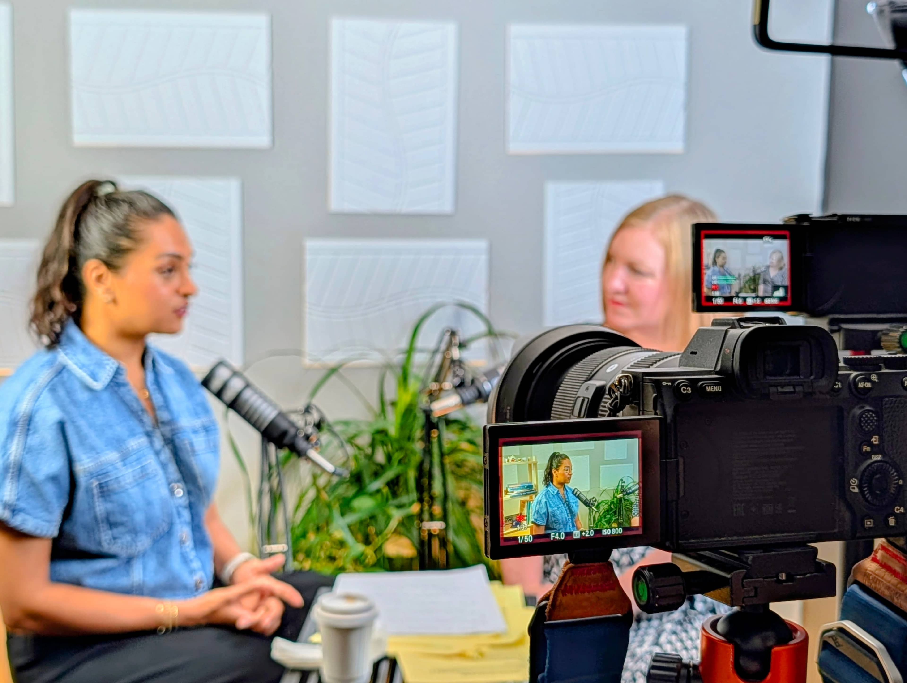 Two women are seated in a recording studio, one speaking into a microphone while the other listens, with cameras capturing the scene from different angles.