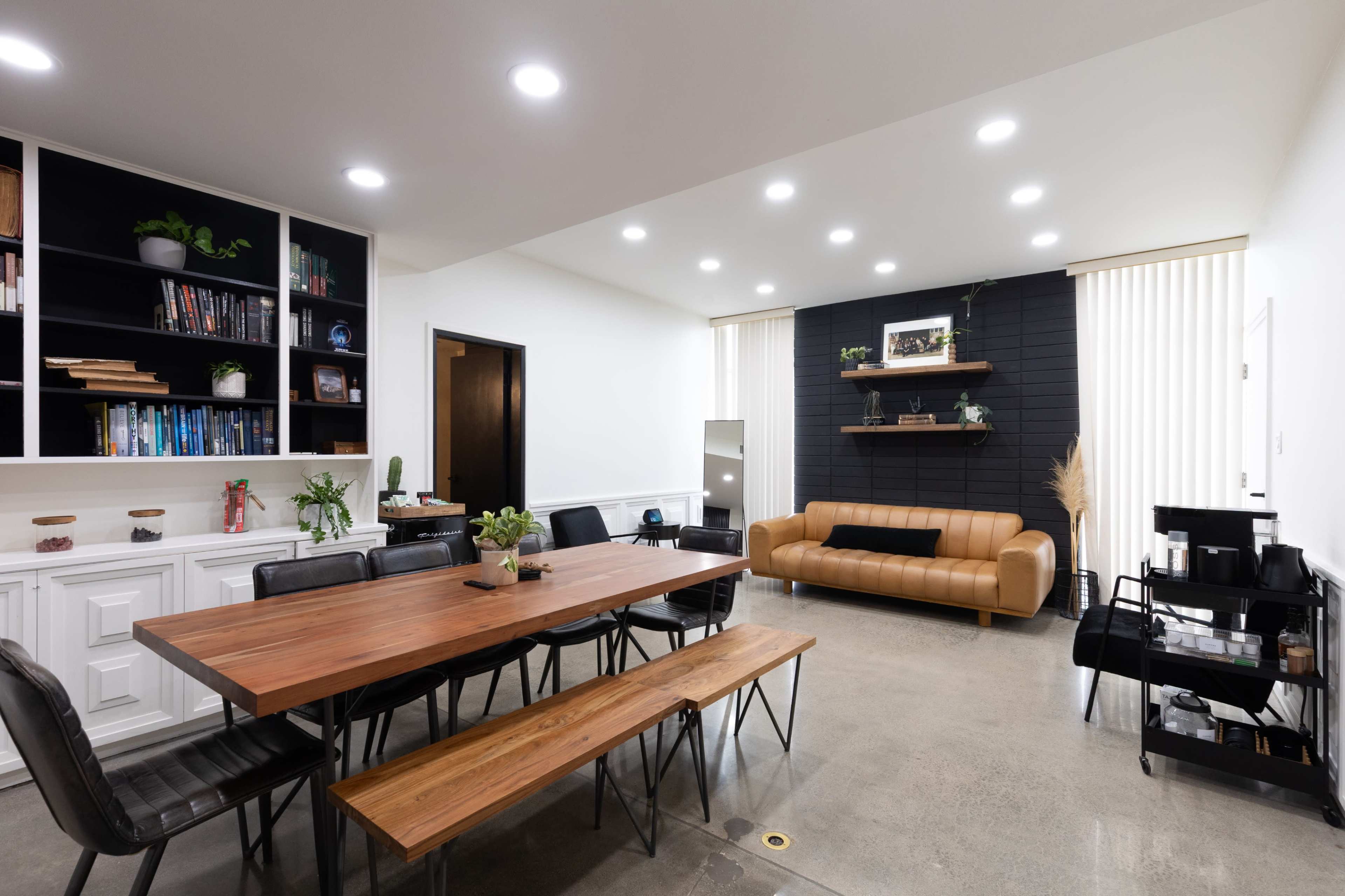 The image shows a modern meeting room featuring a long wooden dining table surrounded by black chairs, a brown leather sofa, shelving with plants and books, and large windows with blinds.