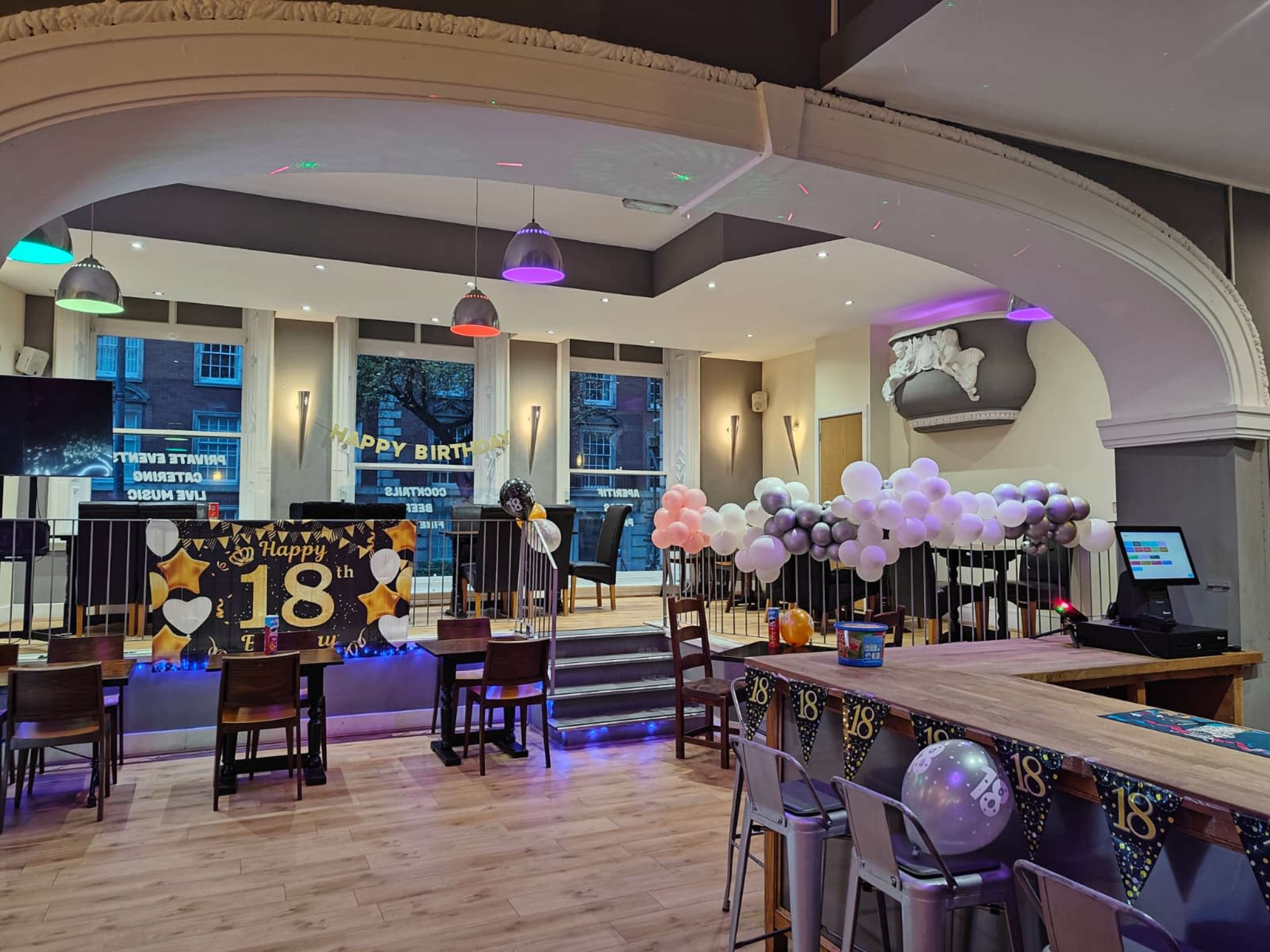 The image shows a decorated indoor space for an 18th birthday celebration, featuring balloons, a banner, and tables set up around a bar area.