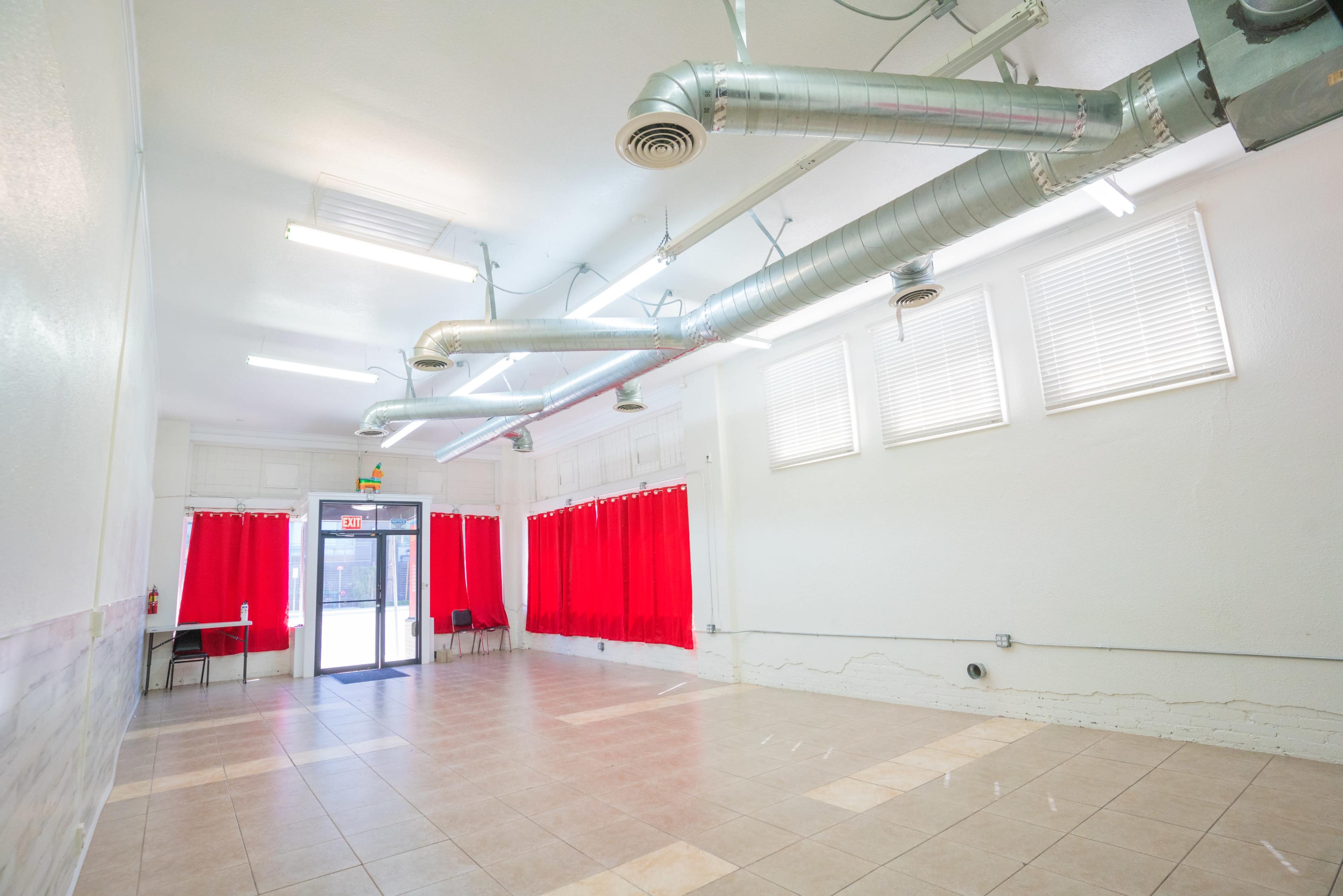 An empty room with tiled flooring, white walls, and large windows covered with red curtains, featuring metal ventilation ducts on the ceiling.