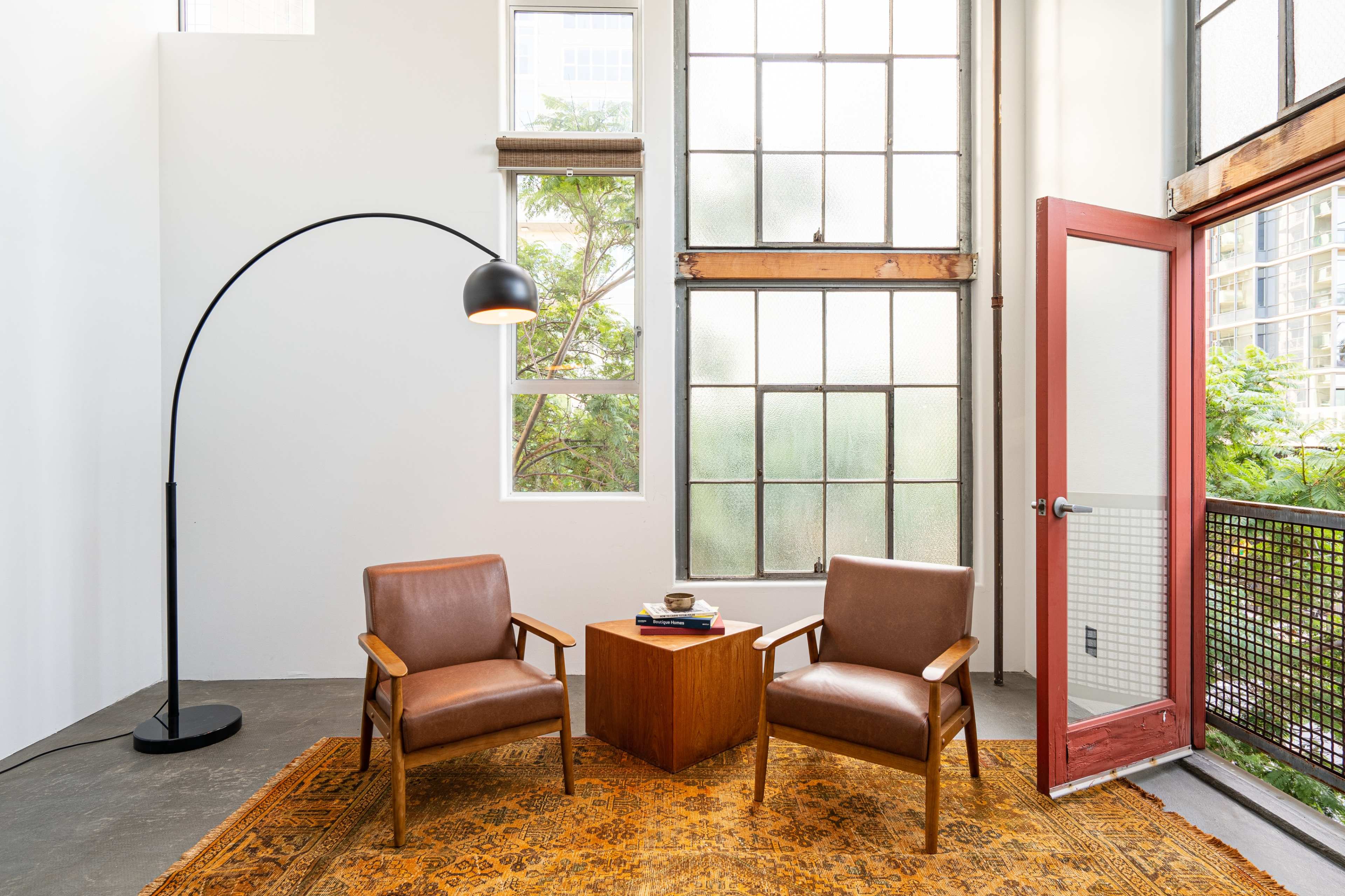 A seating area features two brown chairs beside a wooden side table, illuminated by a floor lamp, with large windows letting in natural light.