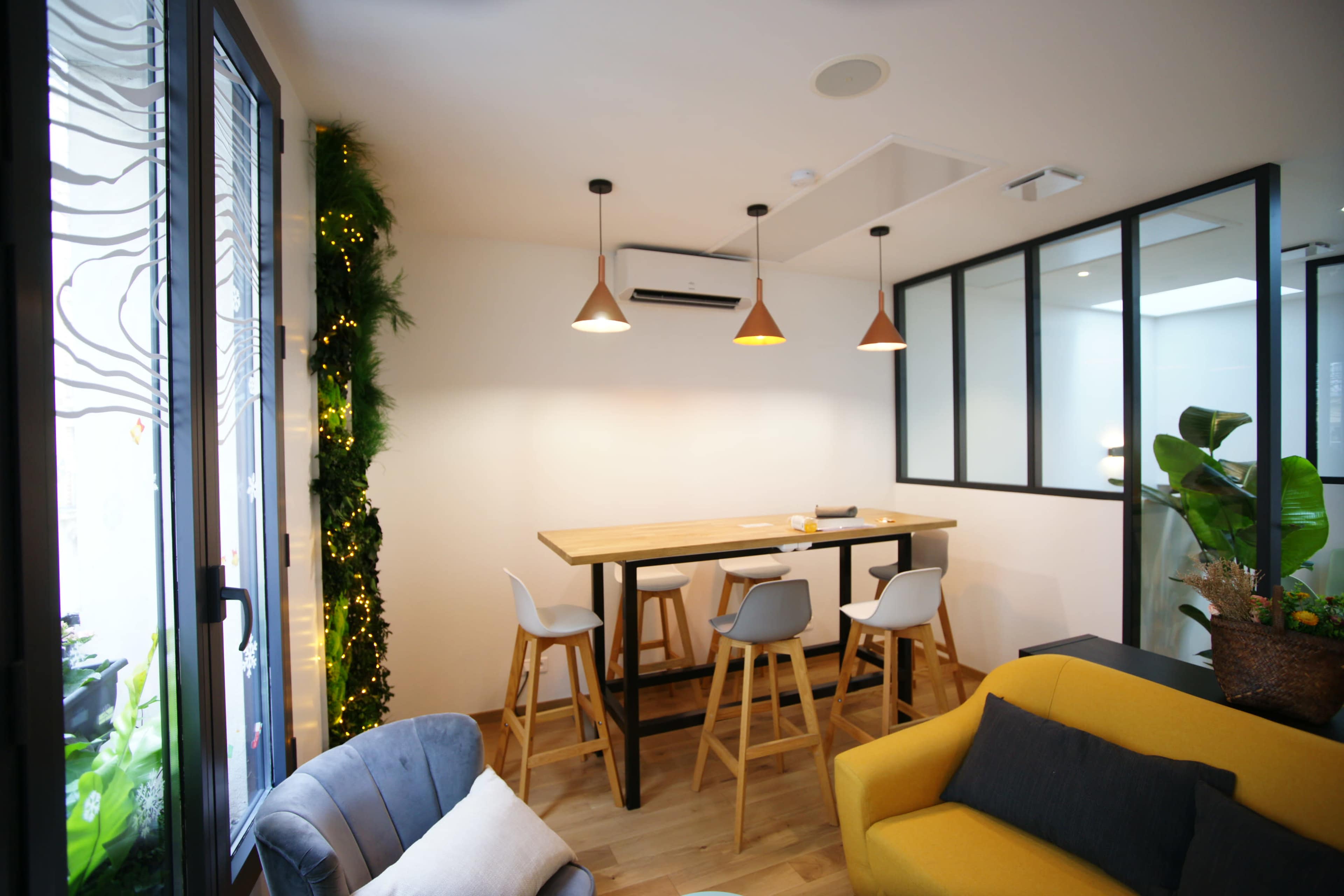 The image shows a modern workspace with a wooden table and high chairs, surrounded by plants and divided by glass walls.