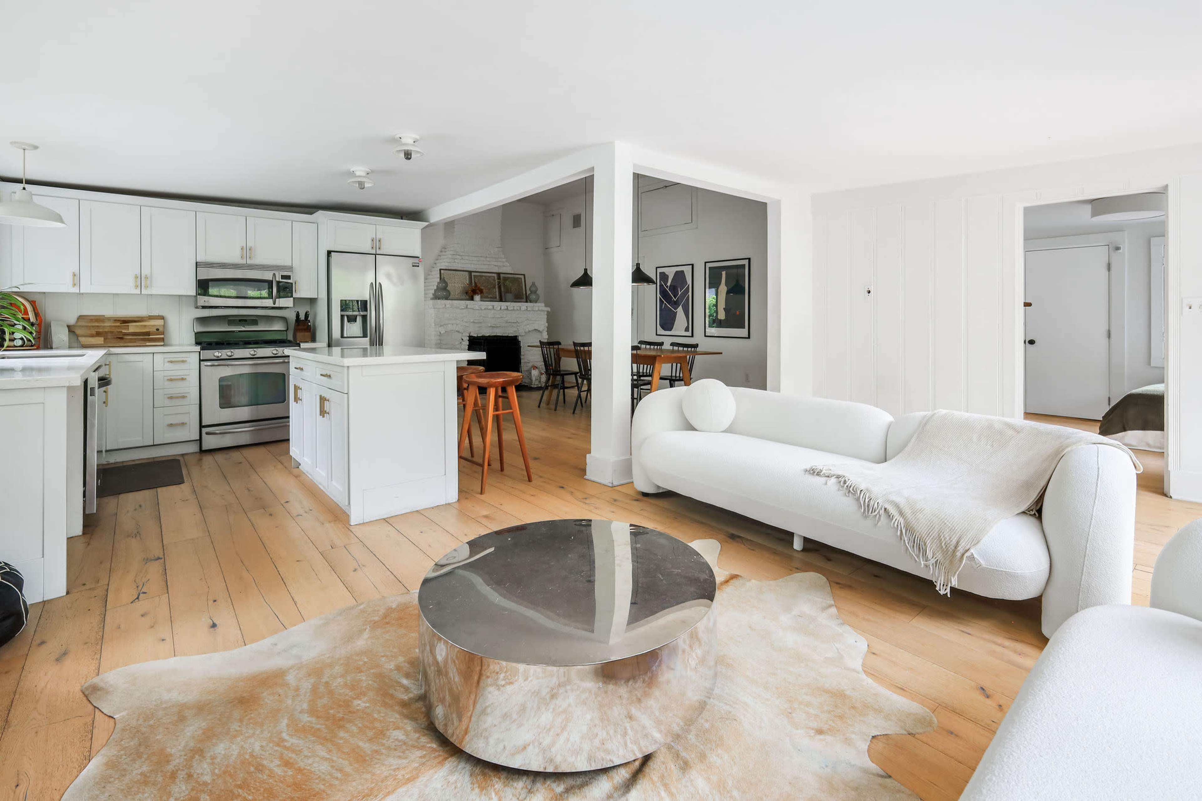 The image shows a modern, open-concept living space featuring a white kitchen, a circular metallic coffee table, and a light-colored sofa on hardwood floors.