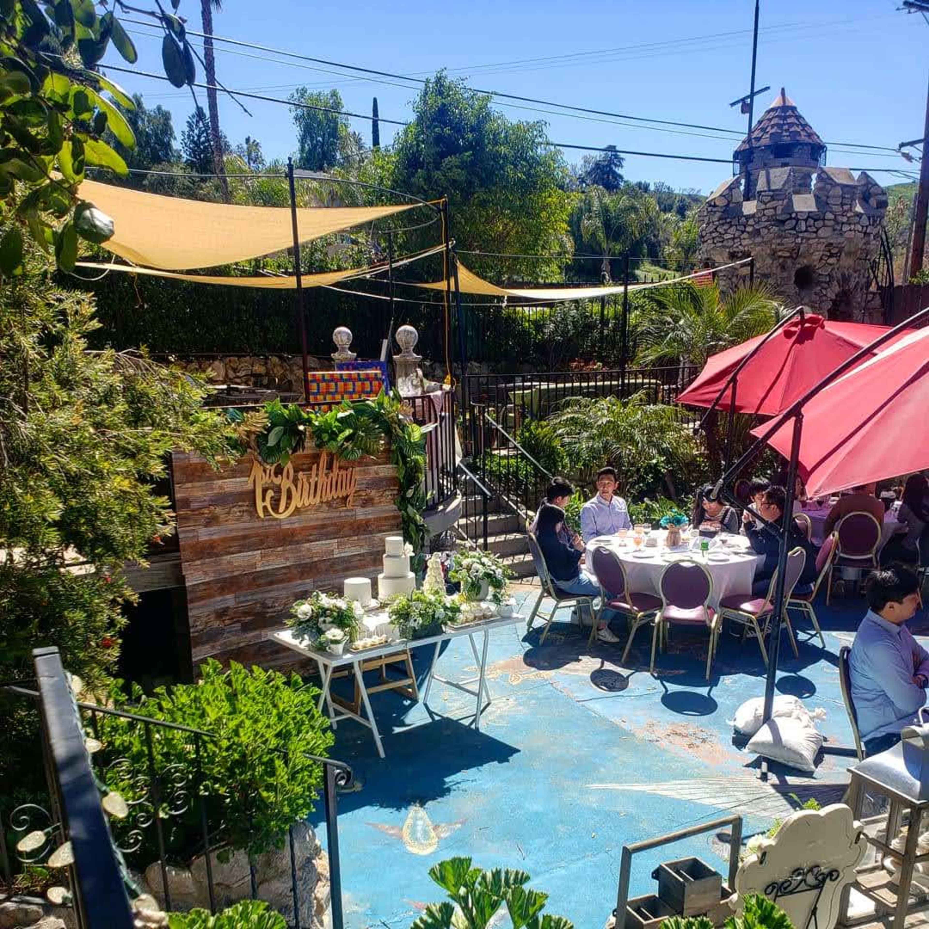 A festive outdoor setting features a decorated table with guests seated around it, surrounded by greenery and a backdrop of a wooden sign.