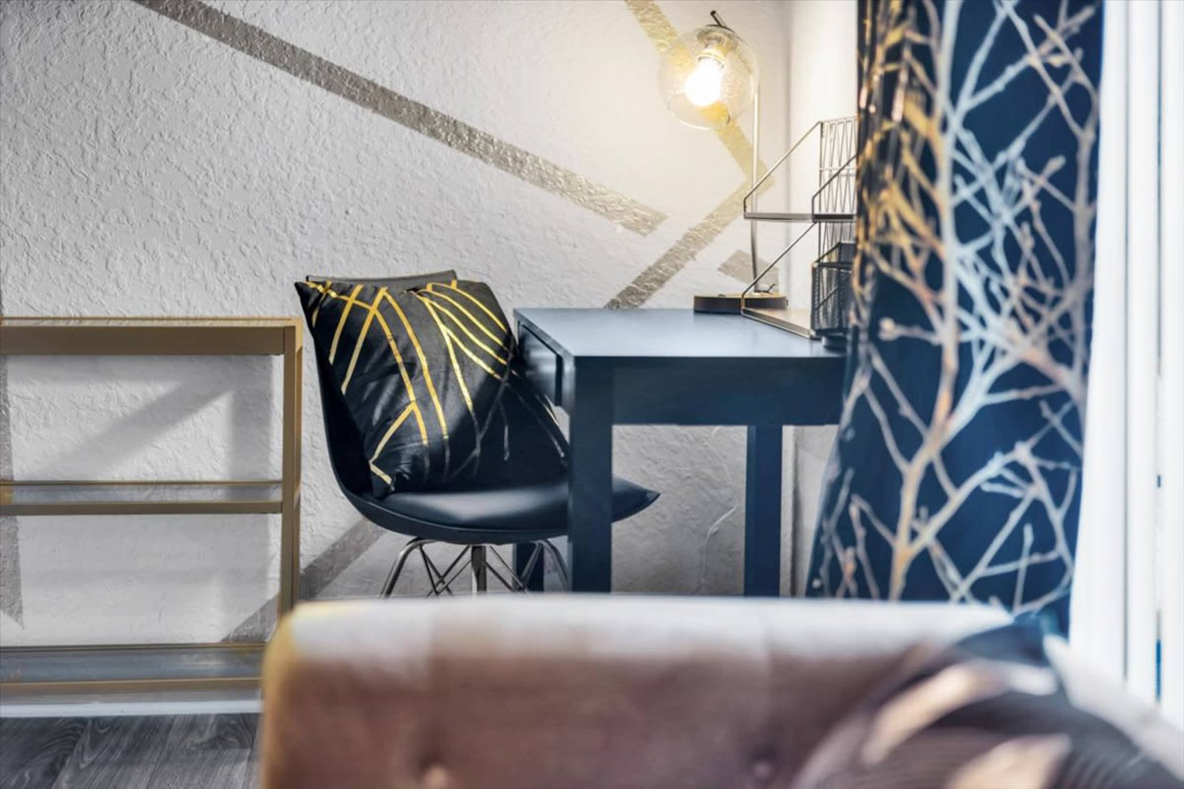 A modern workspace featuring a black desk with a chair, a decorative pillow, and a lamp against a textured wall, alongside an open shelving unit and patterned curtains.