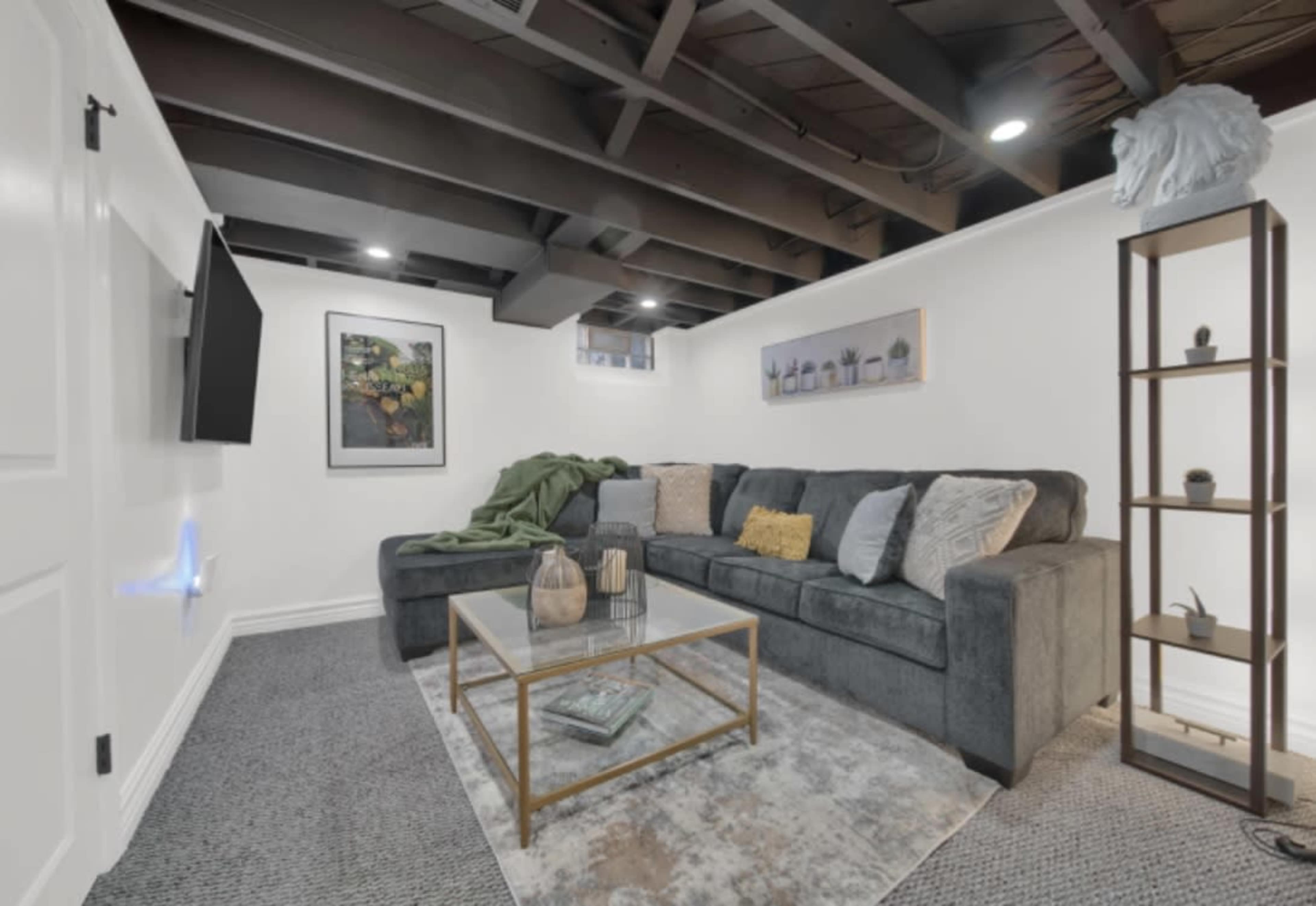 The image shows a modern basement living area featuring a dark gray sectional sofa, a glass coffee table, and decor on a tall shelving unit against a white wall with exposed beams above.