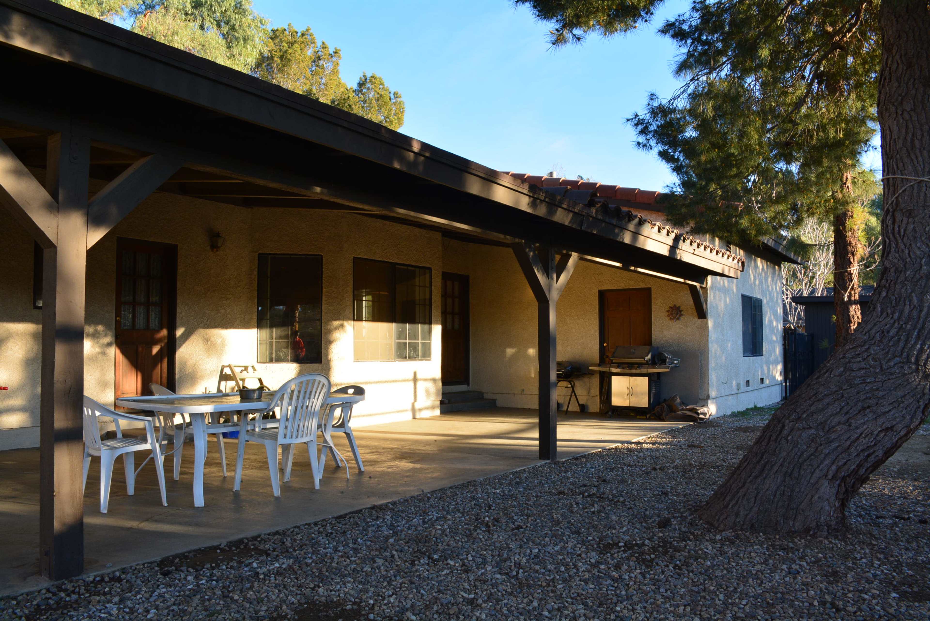 A covered patio with white plastic chairs and a table is adjacent to a single-story house surrounded by trees and gravel.
