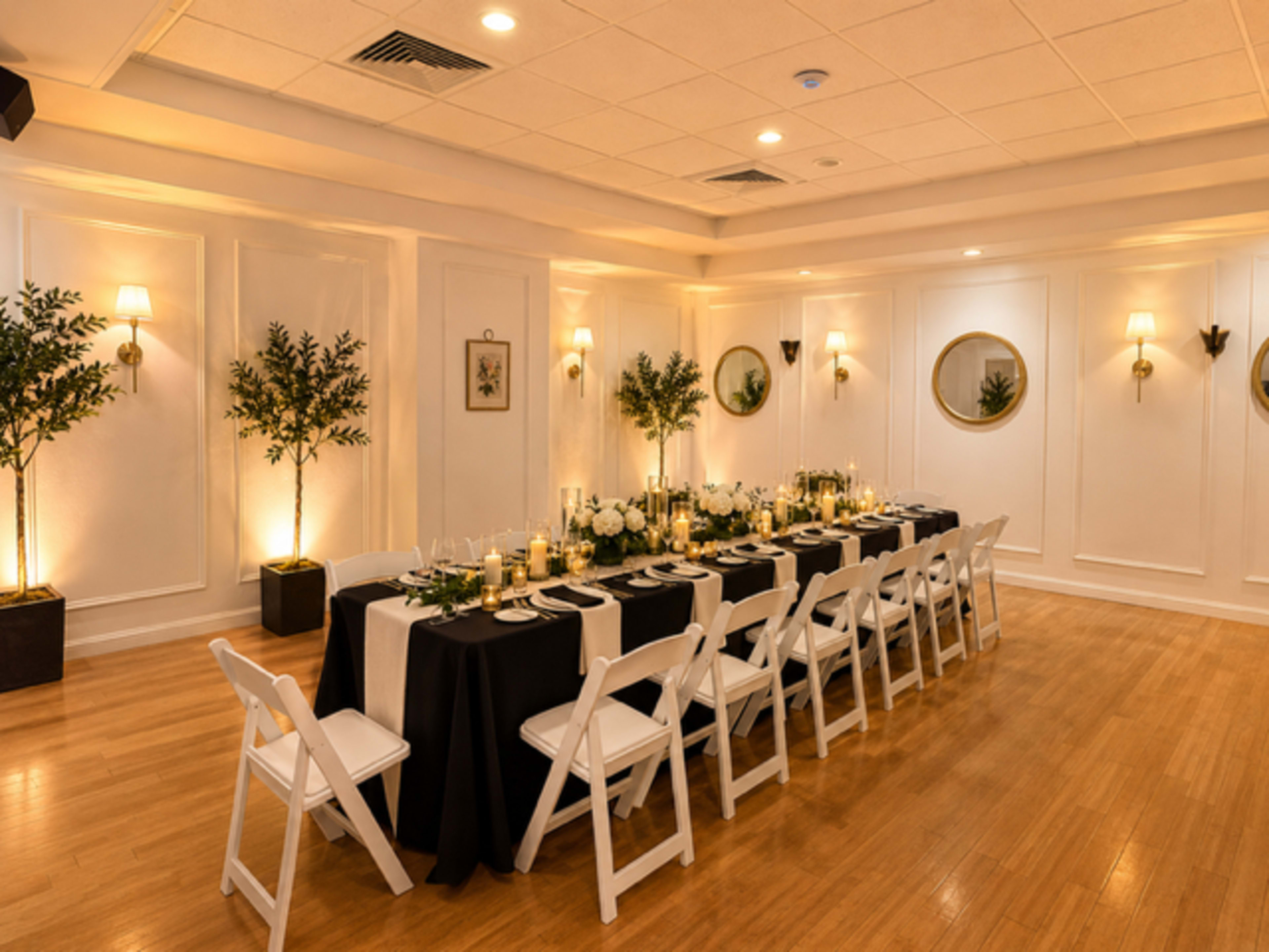 A long, elegantly set dining table with white chairs is arranged in a well-lit room adorned with plants and decorative mirrors.