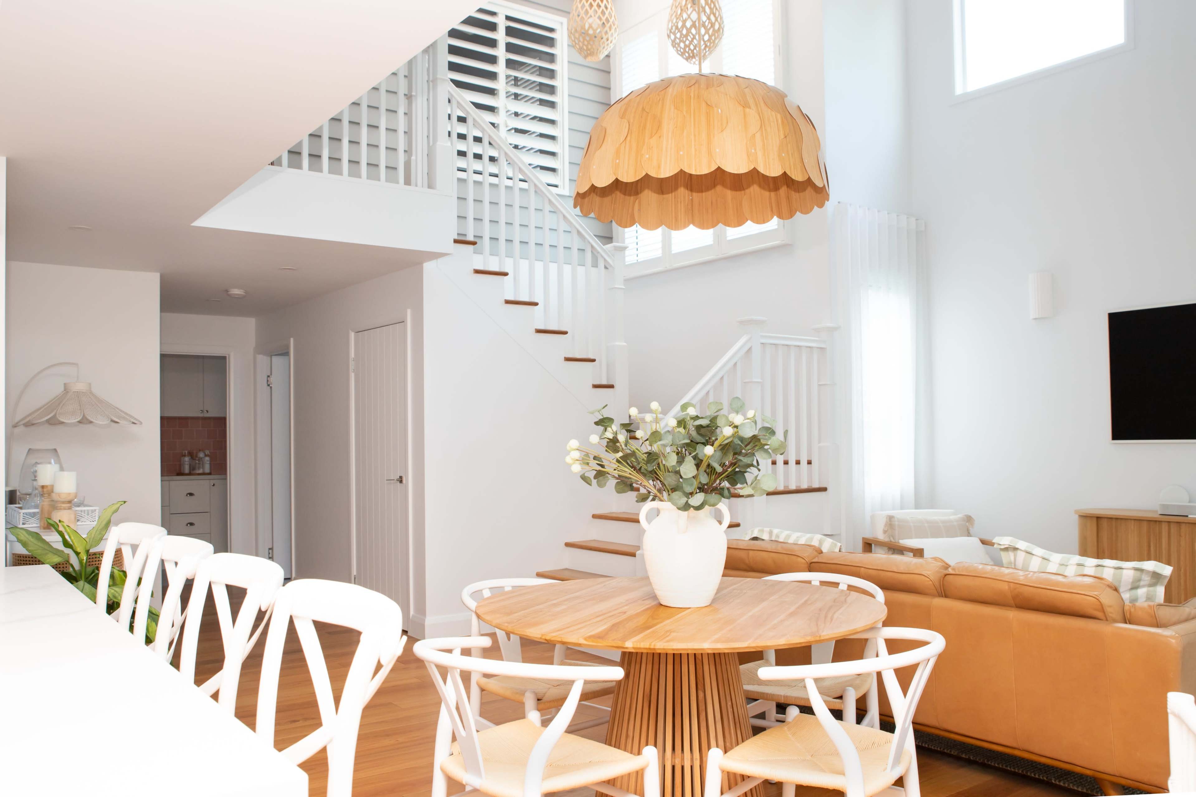 A modern living space features a round wooden dining table with white chairs, a leather sofa, and a staircase, accented by a large pendant light and natural light from high windows.