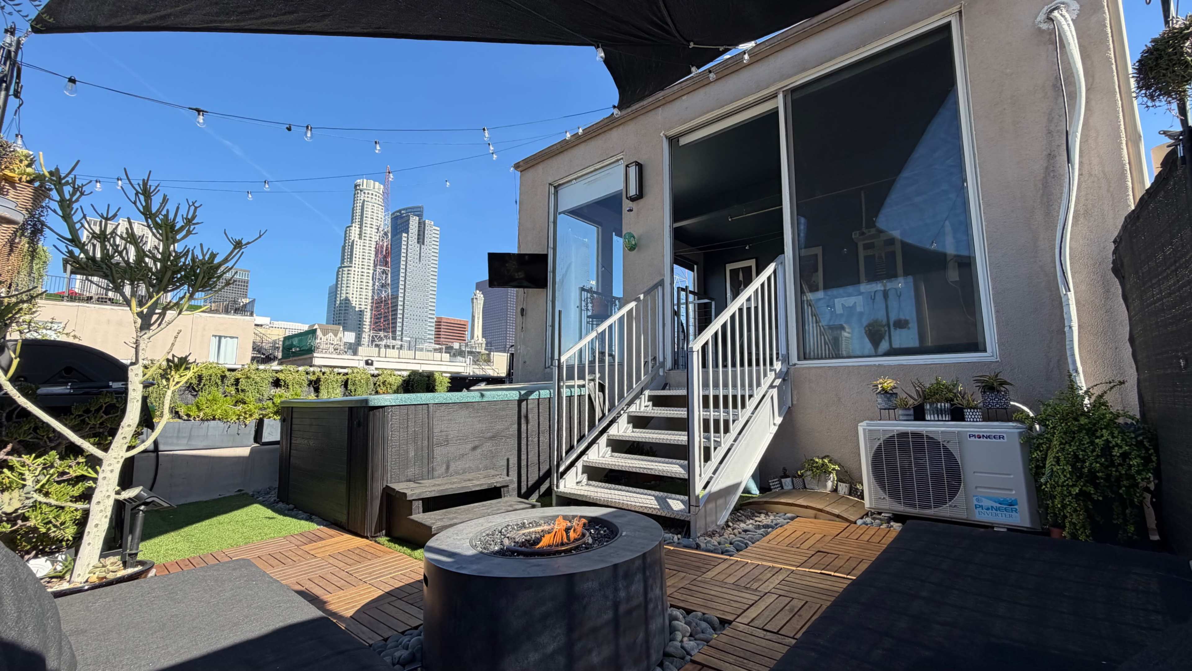 A rooftop space features a fire pit, outdoor seating, and a view of tall buildings under a clear blue sky.