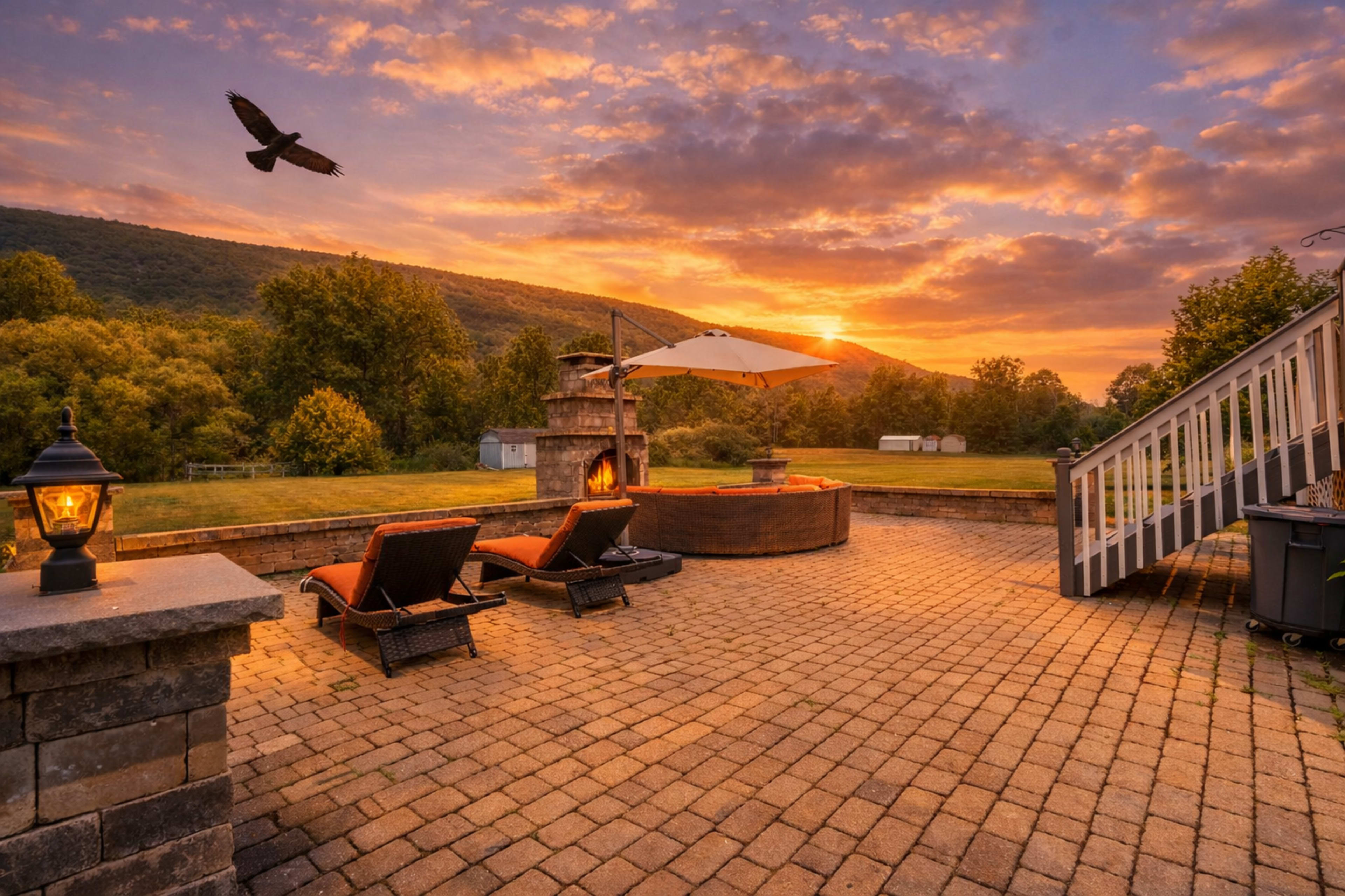 The image shows a patio area with lounge chairs, a fire pit, and an umbrella, set against a vibrant sunset and a mountainous landscape.