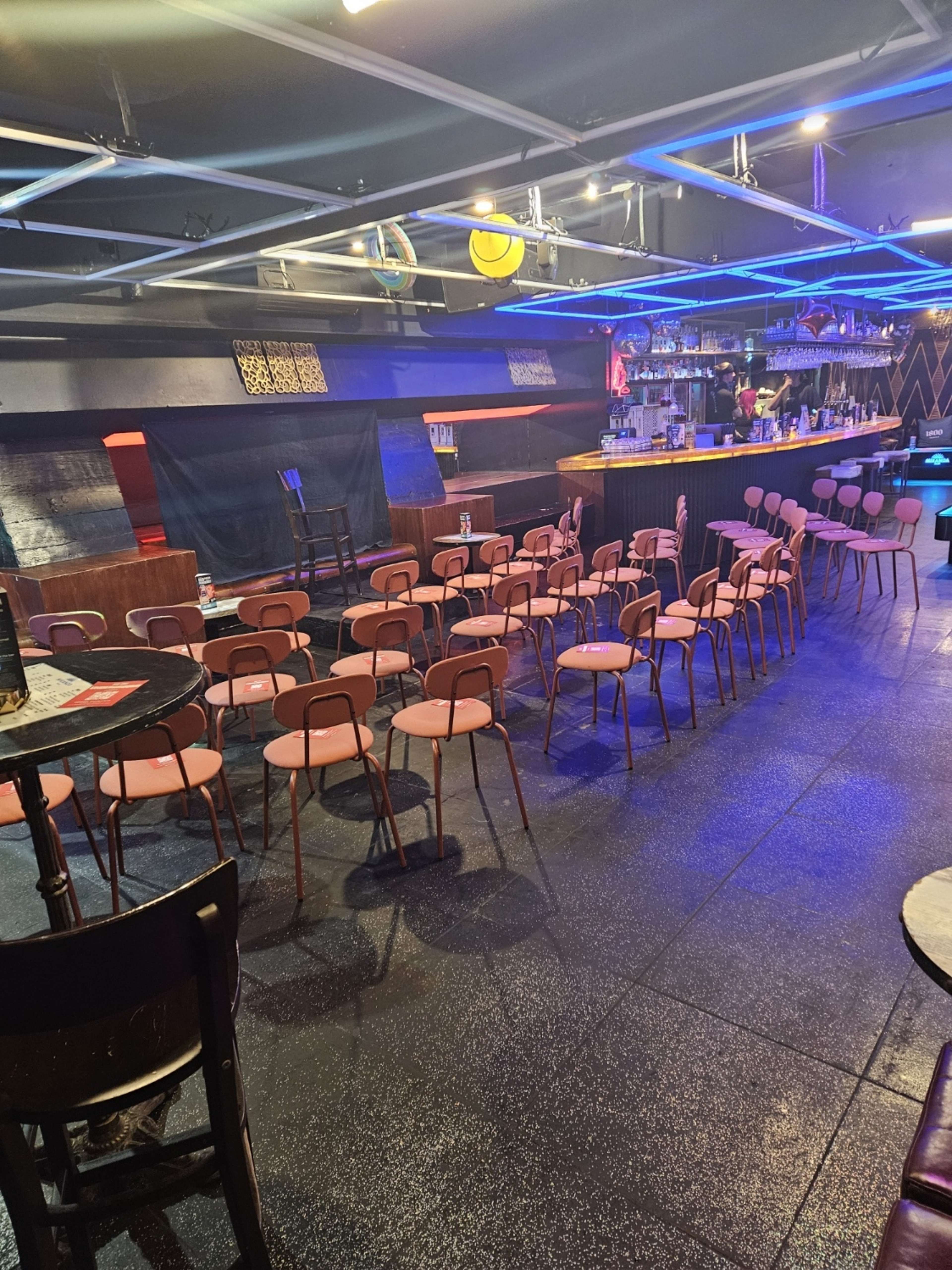 The image shows a dimly lit venue with rows of pink chairs facing a bar and a small stage area.