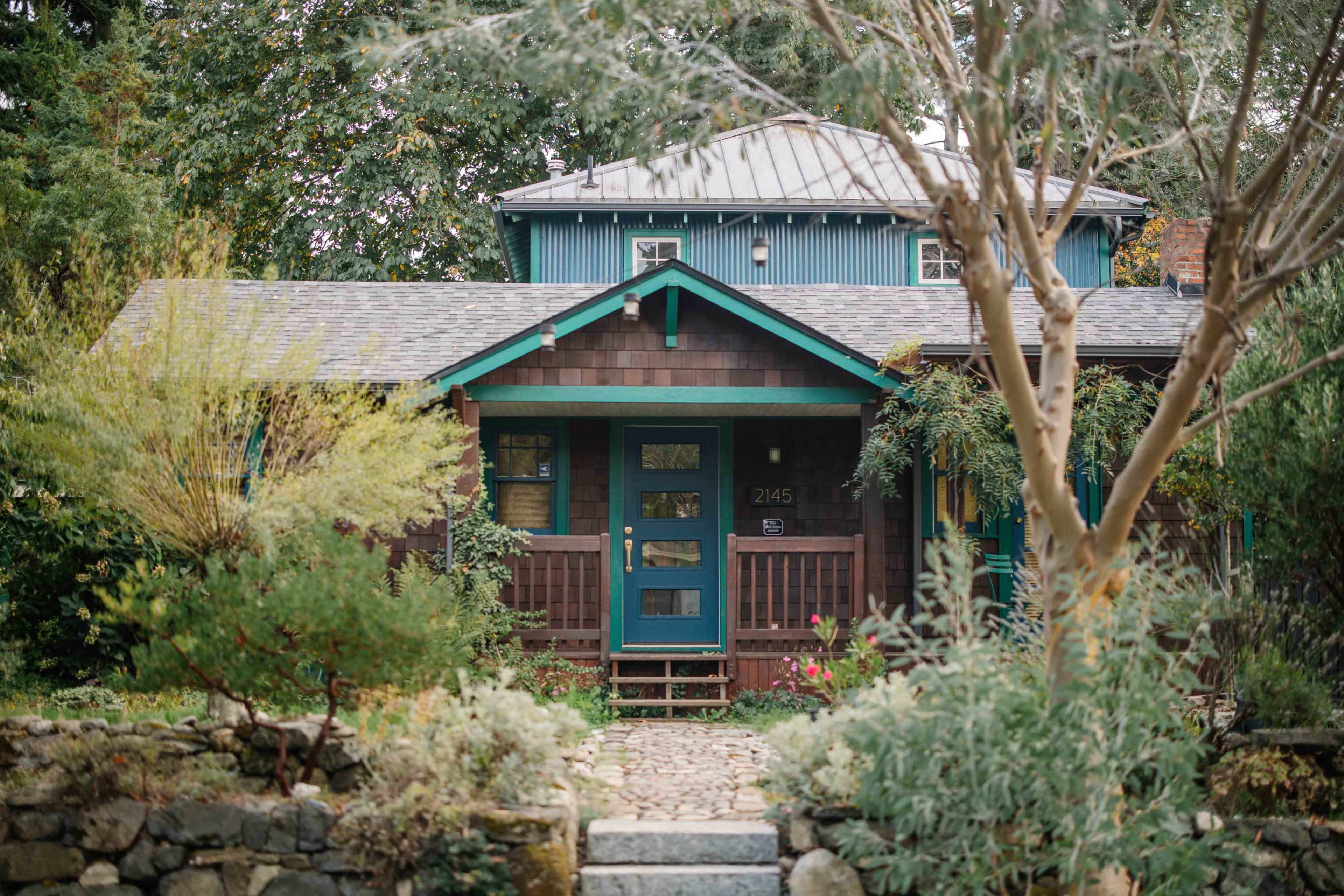 The image shows a rustic, two-story house with a blue roof, green trim, and a front porch surrounded by lush greenery and stone pathways.